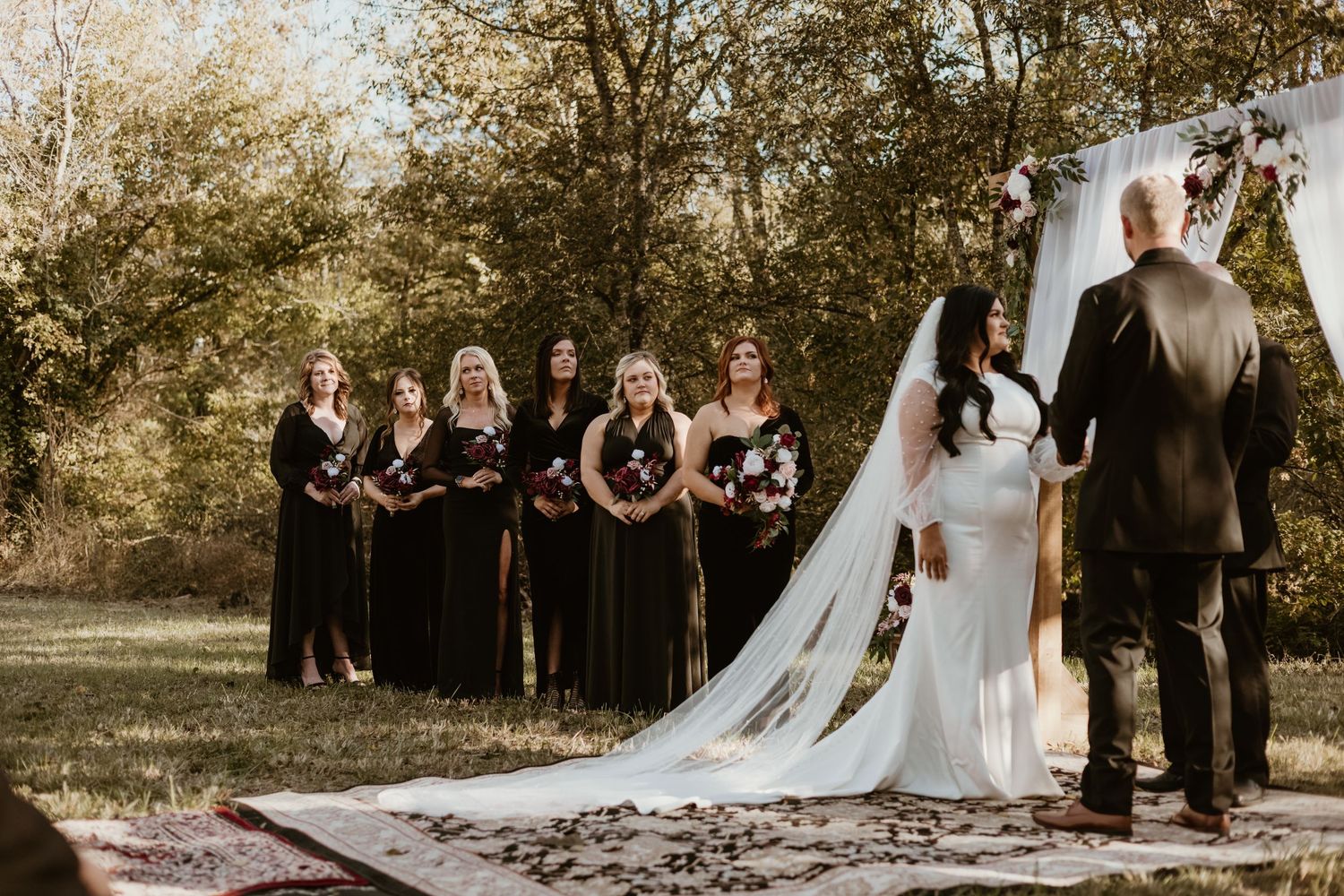 A bride and groom stand at the altar with bridesmaids in black dresses during an outdoor autumn wedding ceremony.