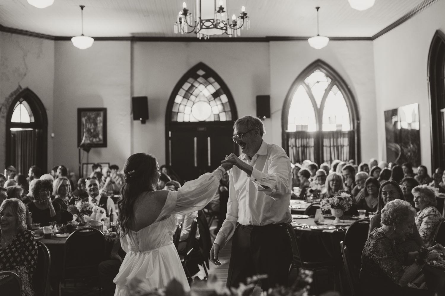 A couple dances during their wedding reception in a historic church hall filled with celebrating guests.