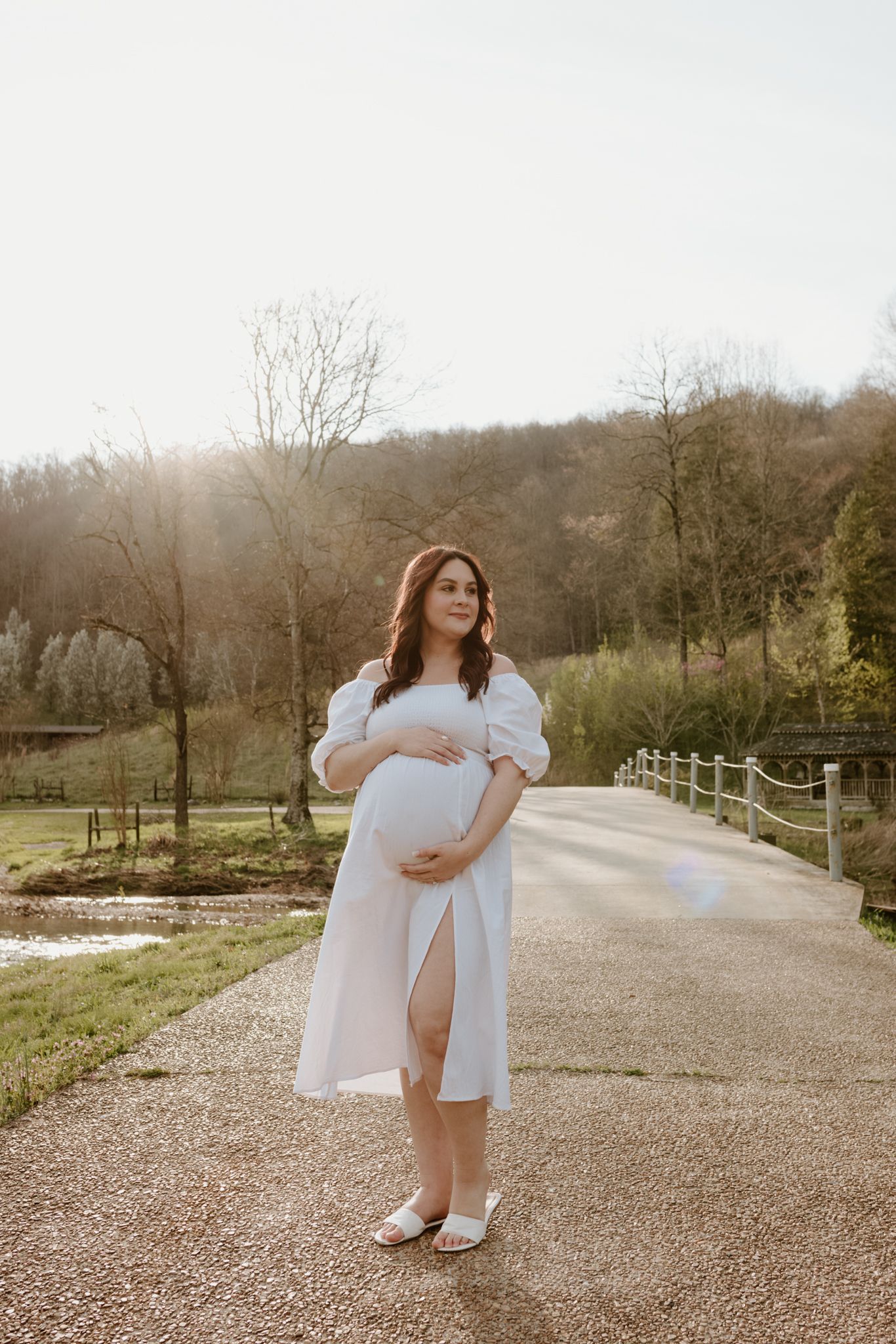 Expectant mother in a white dress poses on a gravel path with white fence and trees in the background.