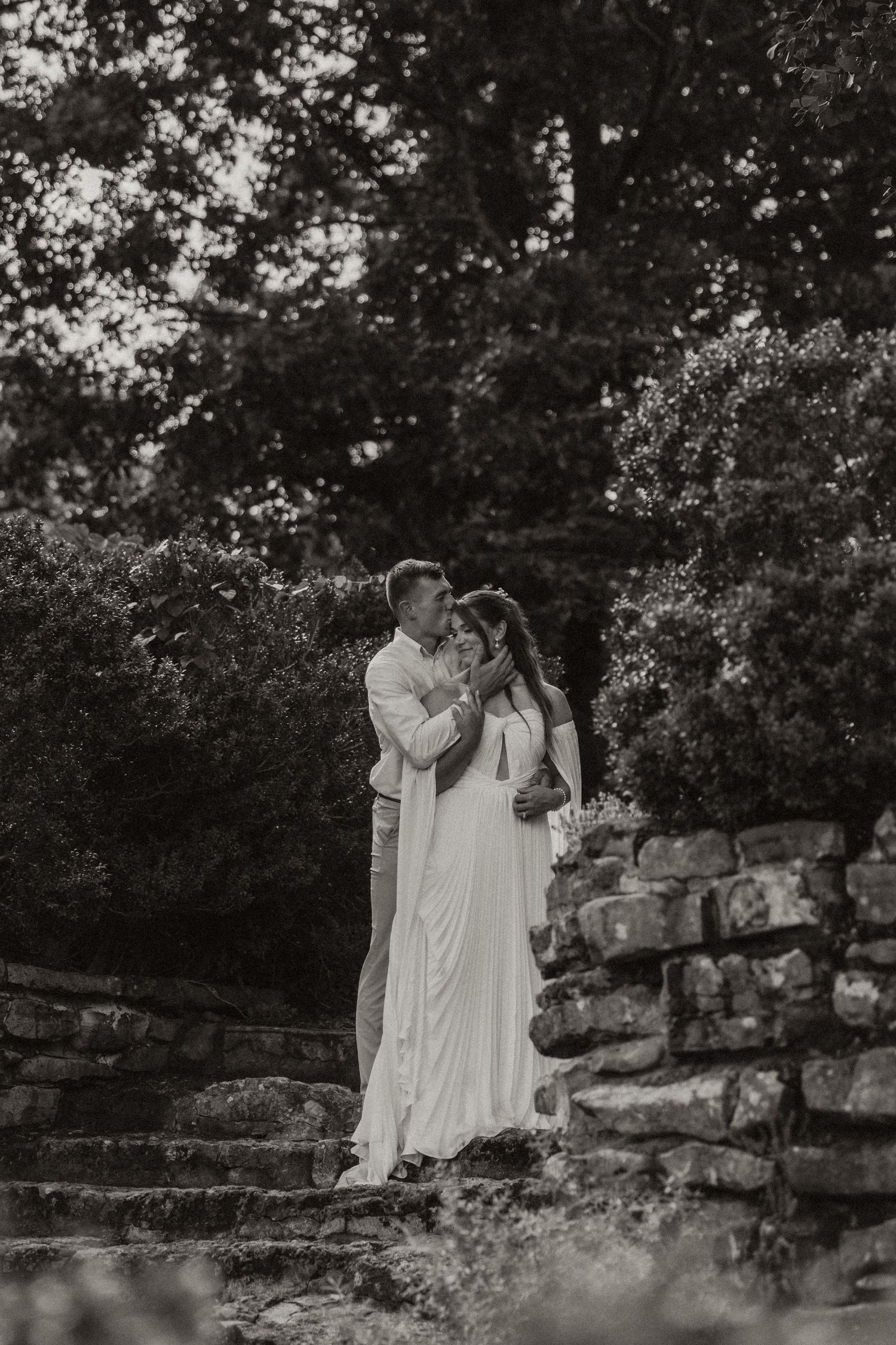 Black and white photo of a couple embracing on stone steps surrounded by lush greenery, bride in elegant white gown.