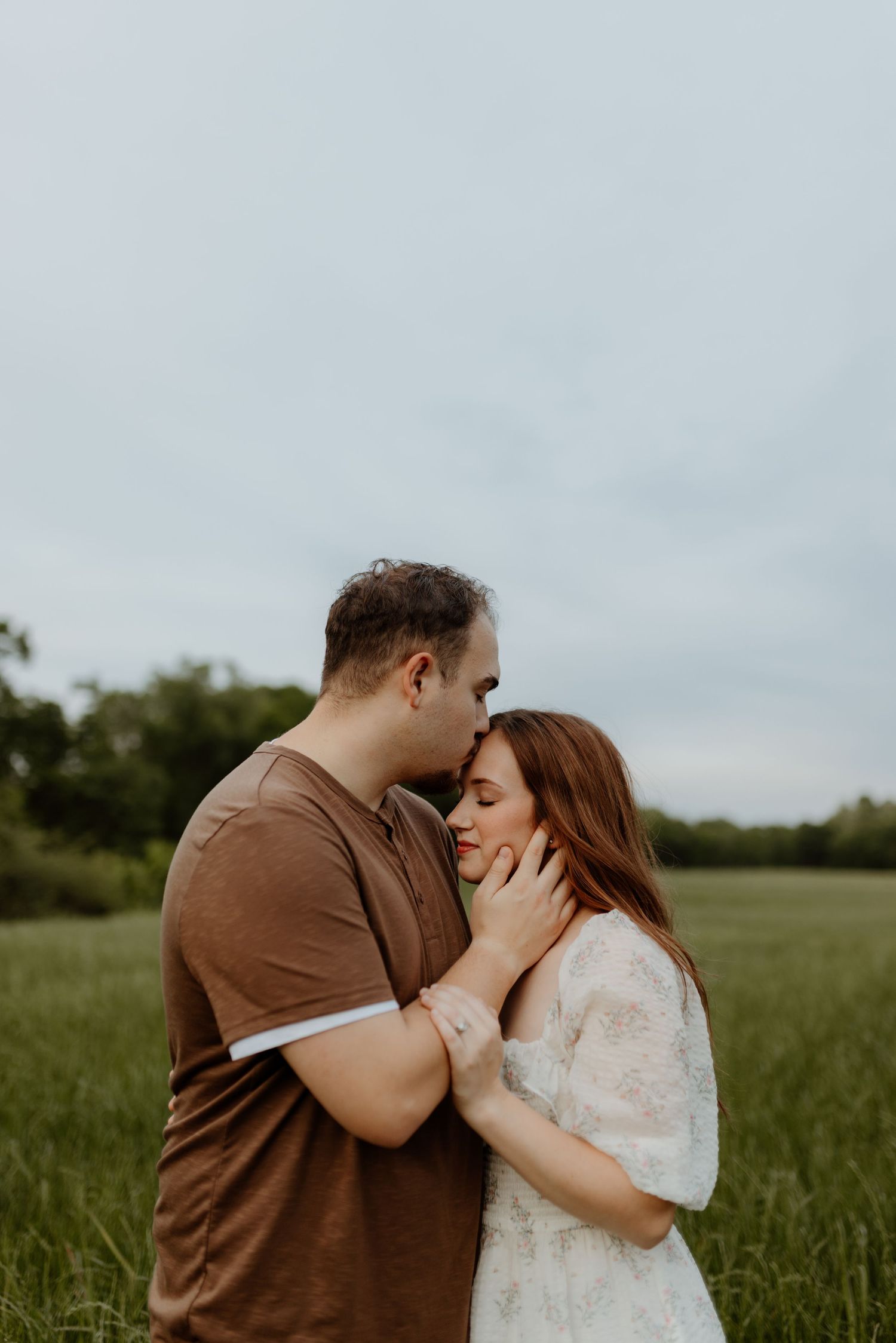A couple shares a tender moment in a green field, the man gently cradling the woman's face as they lean in close.