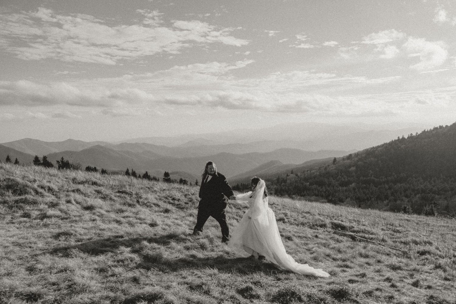 Couple twirling in wedding attire on hilltop with stunning mountain landscape in black and white.