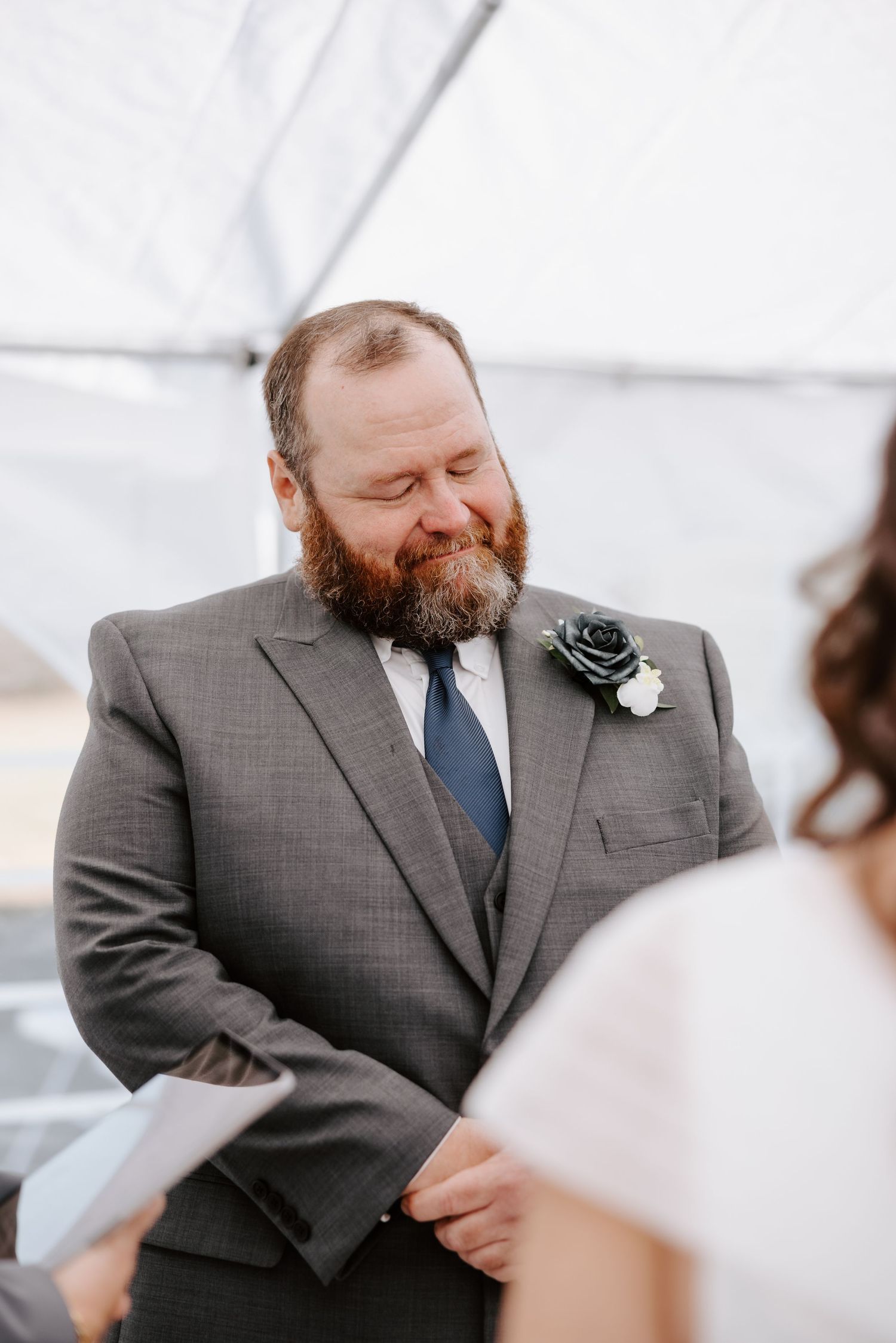 Groom in gray suit with navy boutonniere smiling emotionally during outdoor wedding ceremony under white tent.