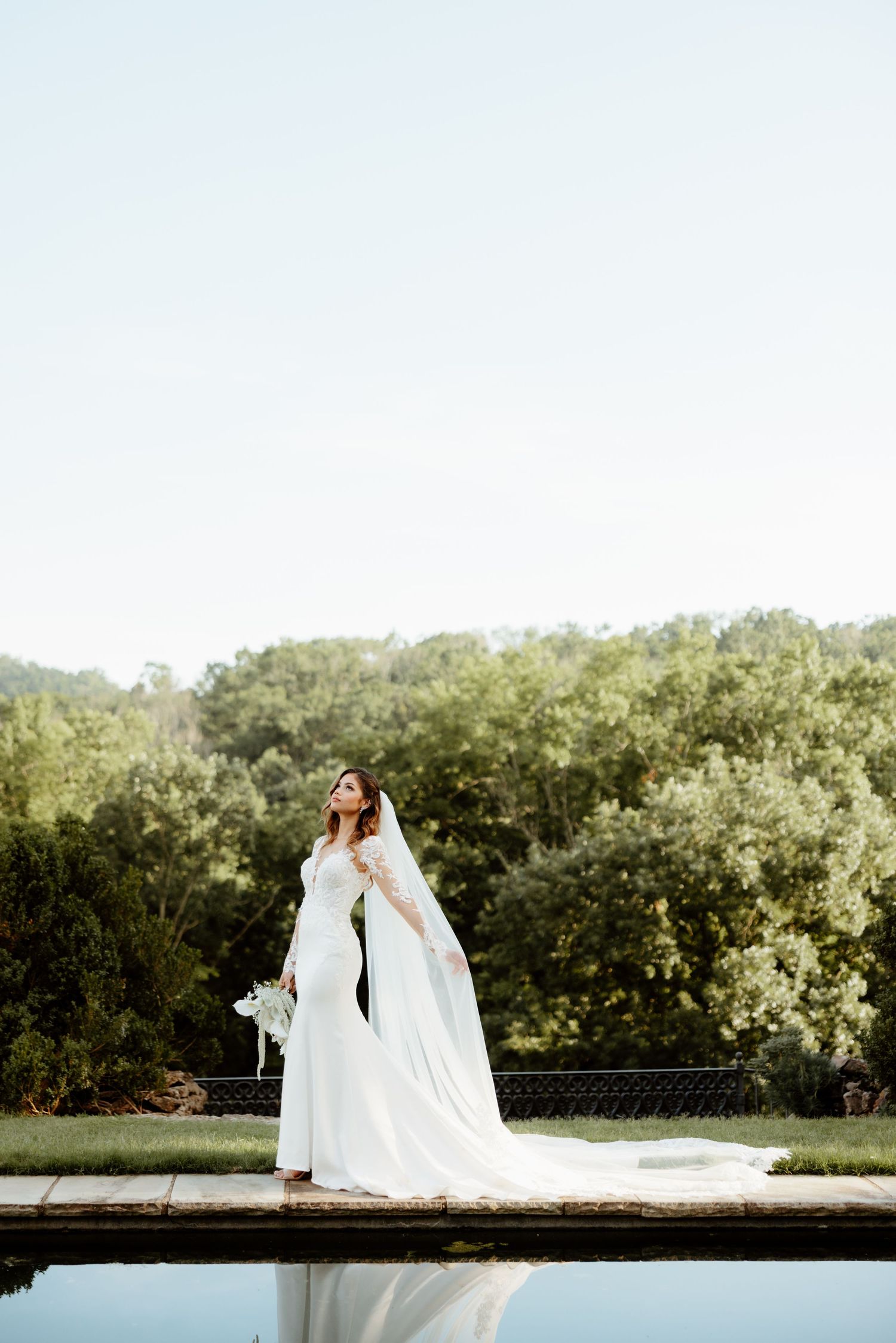 Bride in white gown and cape veil stands by reflective pool with lush green trees backdrop on wedding day.