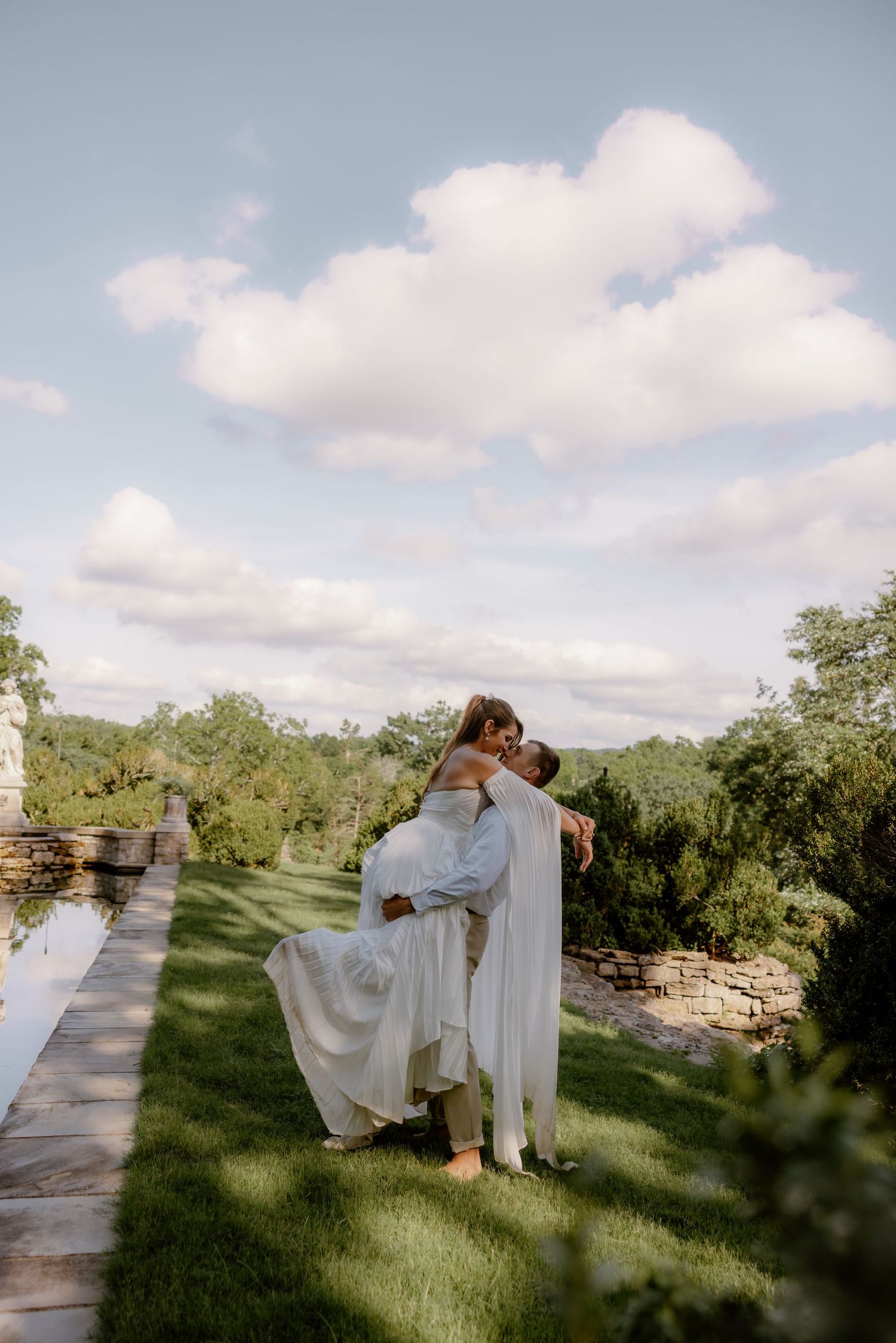 Groom lifting bride in white gown outdoors on lush green lawn with scenic garden backdrop and cloudy blue sky.