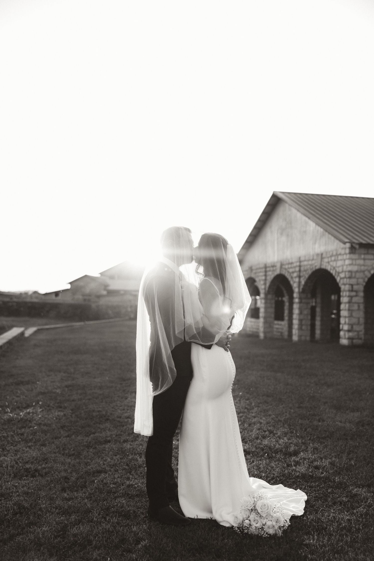 Artistic black and white wedding portrait with sun flare against historic stone architecture.