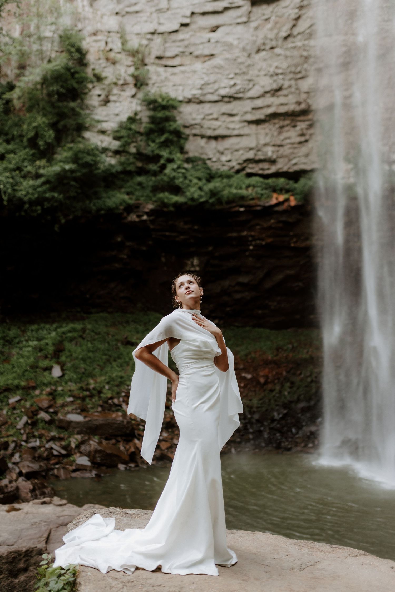 Elegant bride in a white cape gown poses gracefully in front of a stunning waterfall surrounded by rocky cliffs.
