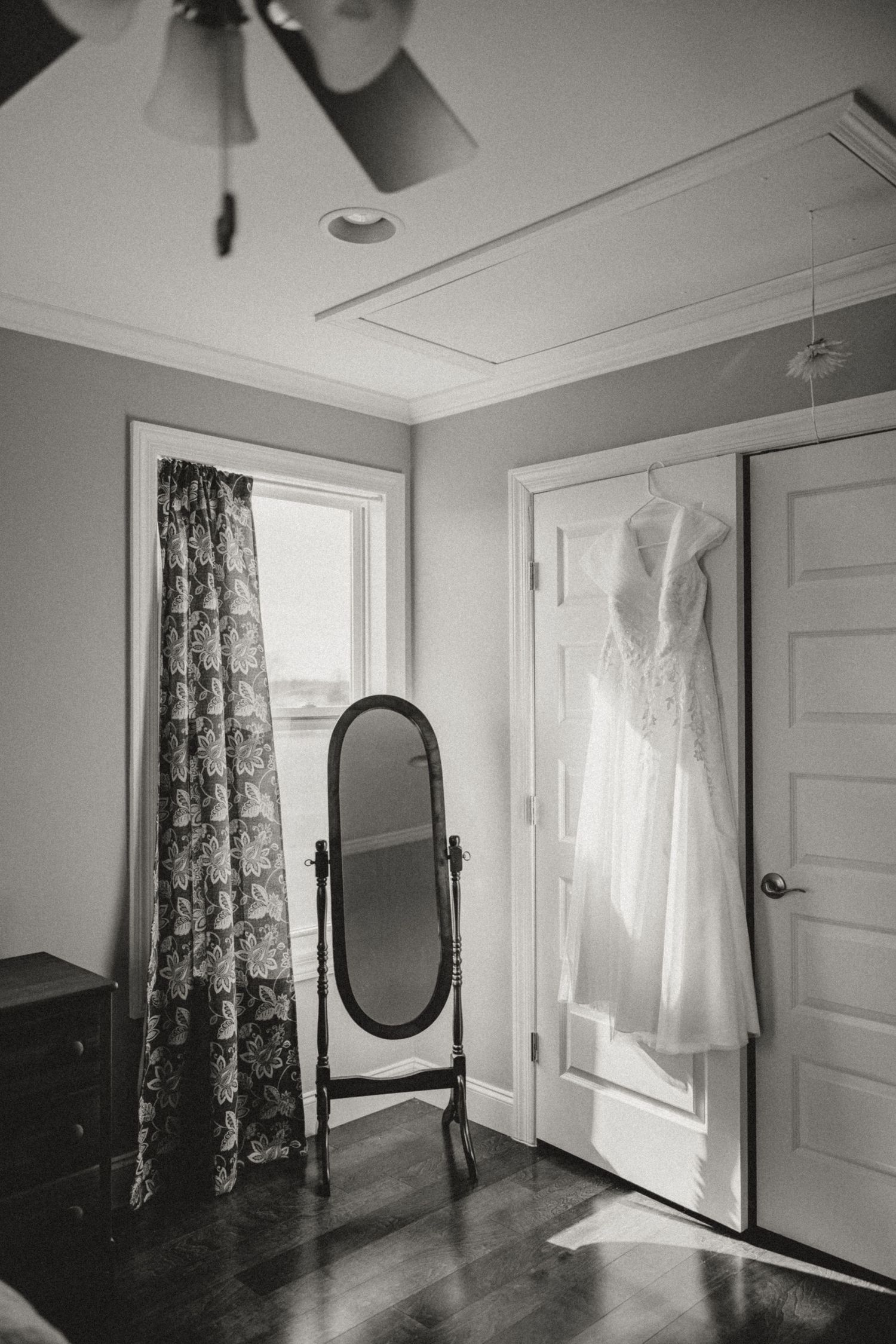Black and white photo of a wedding dress hanging on a door beside a vintage floor mirror in an elegant room.