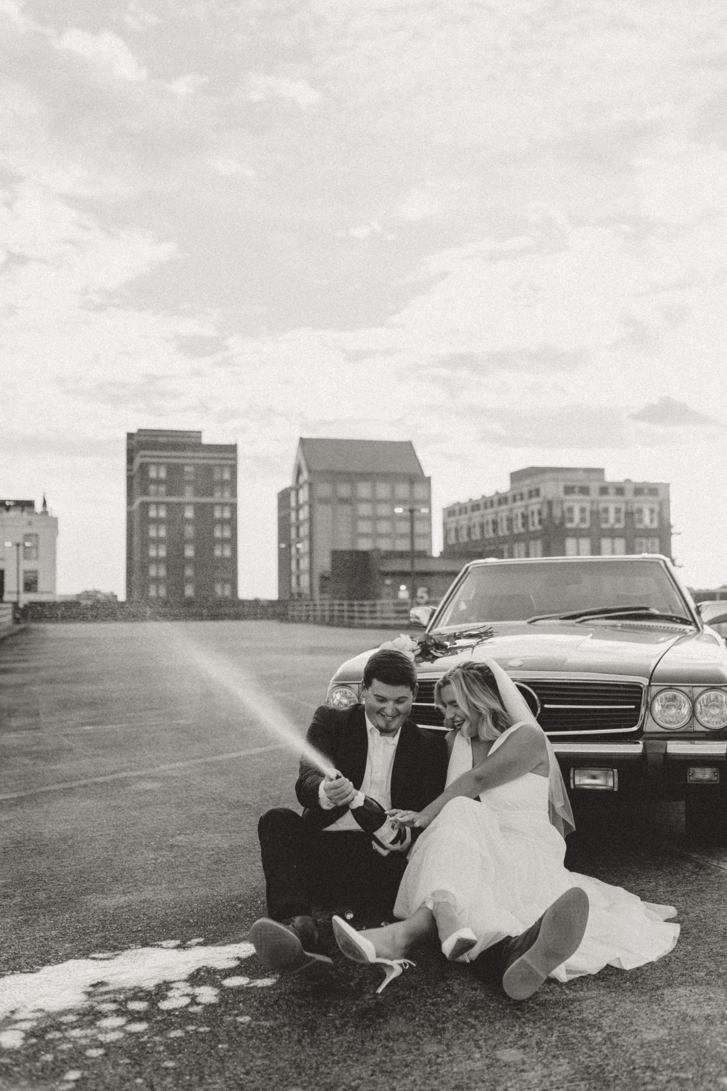 Black and white photo of a couple sitting together on grass near a vintage car with urban buildings in the background.