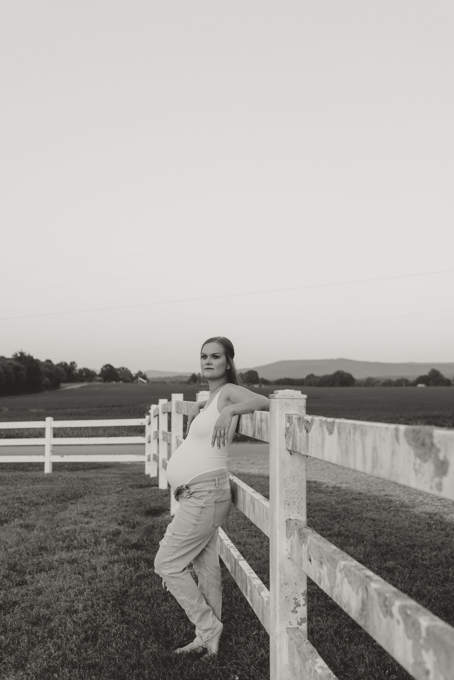 A black and white photo of someone leaning against a white fence in a rural setting.