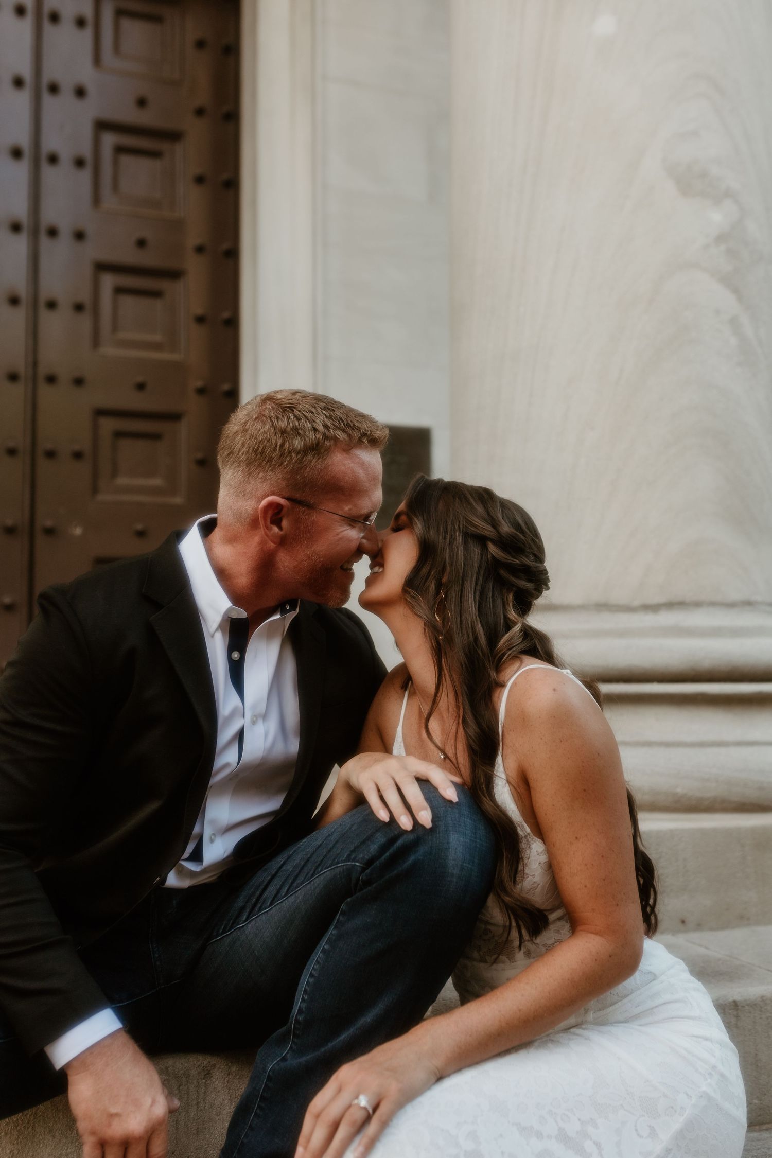 A romantic wedding couple shares intimate kisses sitting against a marble column wall in black and white formal attire.