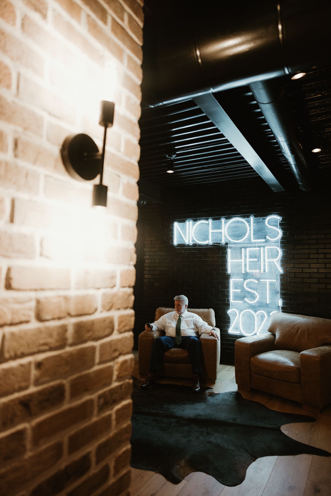 A modern brick wall interior with neon signage reading 'NICHOLS HEIR EST 2022' and leather seating.