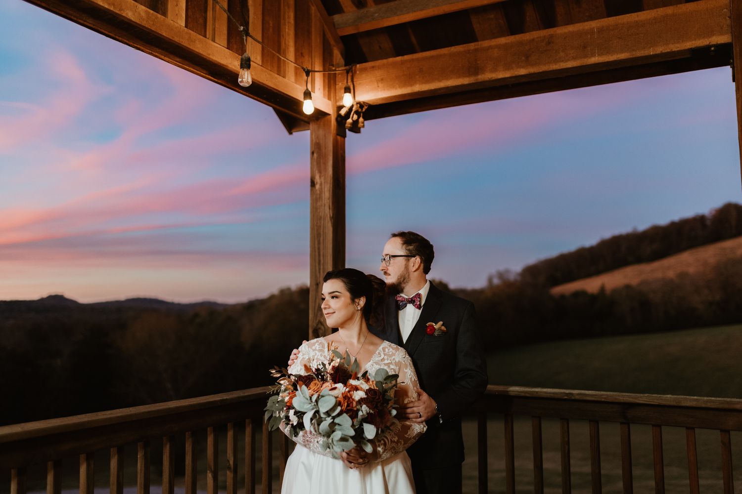 Romantic sunset wedding venue with wooden beams and railings silhouetted against pink and purple sky.