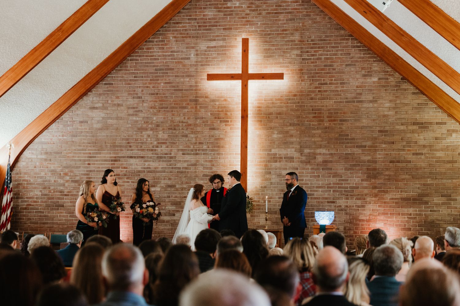 A wedding ceremony takes place in a church sanctuary with exposed brick walls and a large illuminated cross.