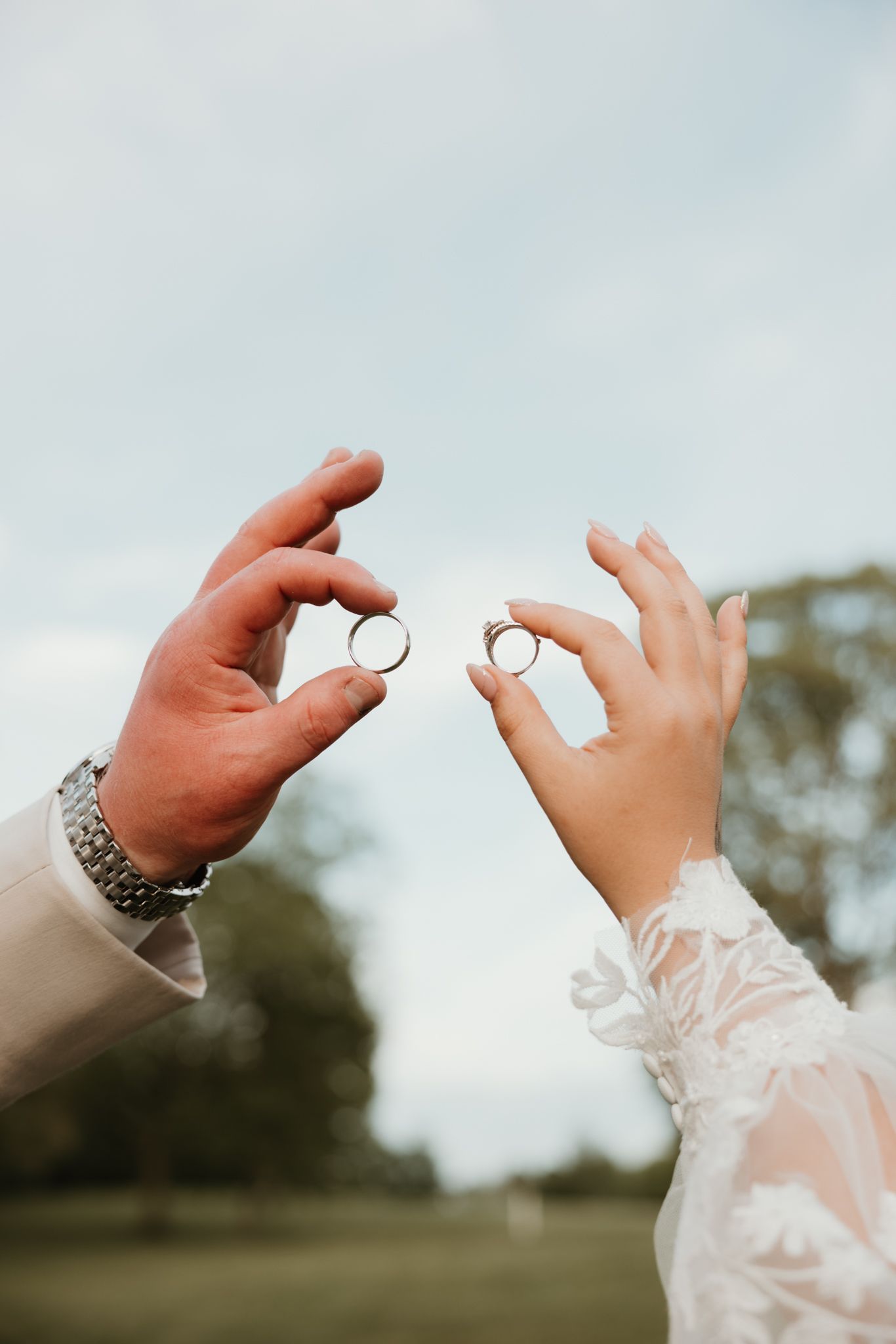 Close-up photograph of two hands exchanging wedding rings against a soft, blurred outdoor background.