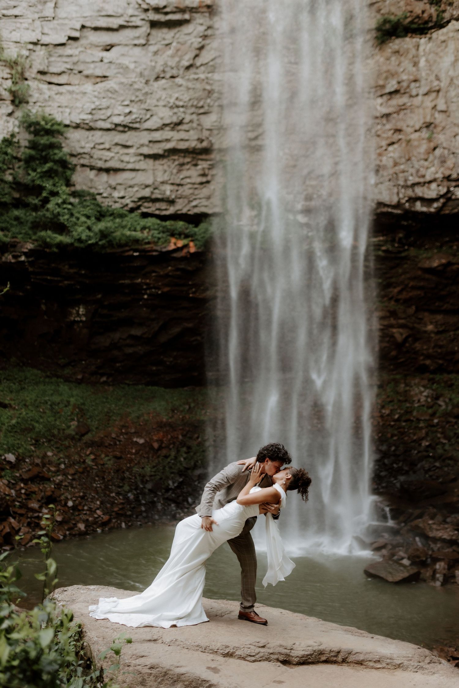 Bride and groom share a romantic dip kiss in front of a stunning waterfall on their wedding day.