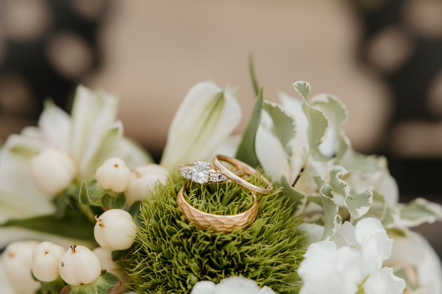 Gold wedding bands and diamond engagement ring nestled in green moss among white flowers and berries.