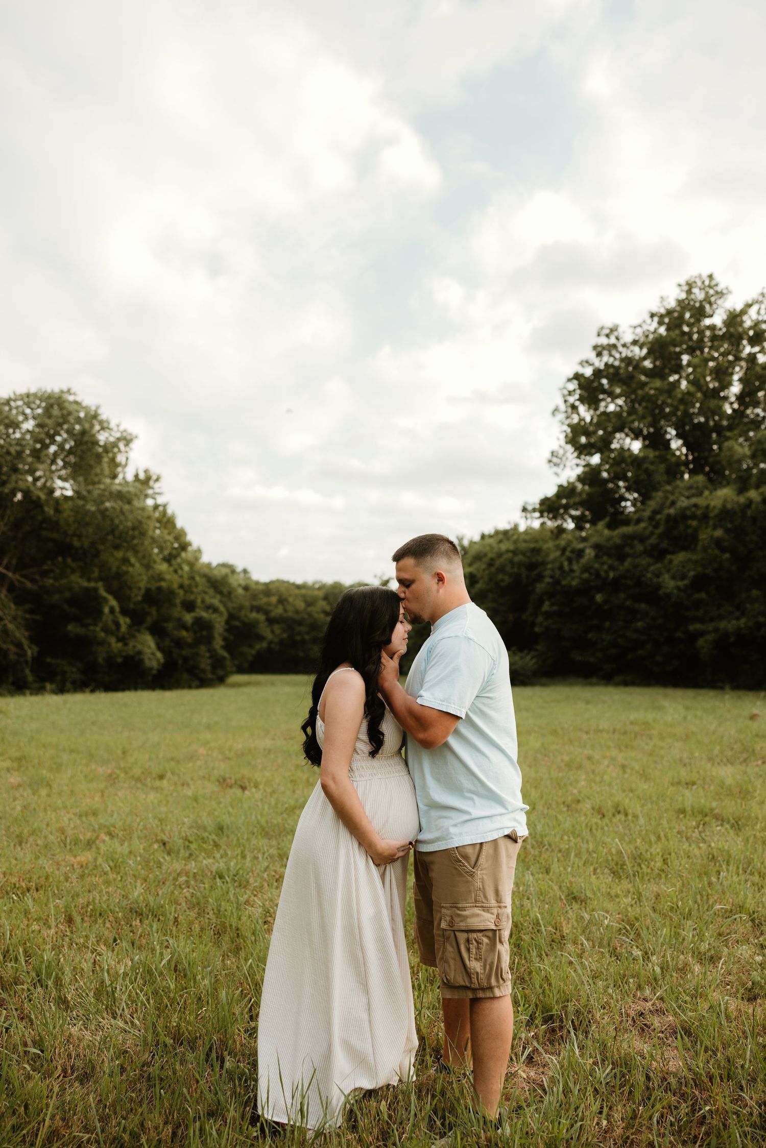 A couple shares a tender moment while standing together in a sunlit grassy meadow with trees in the background.