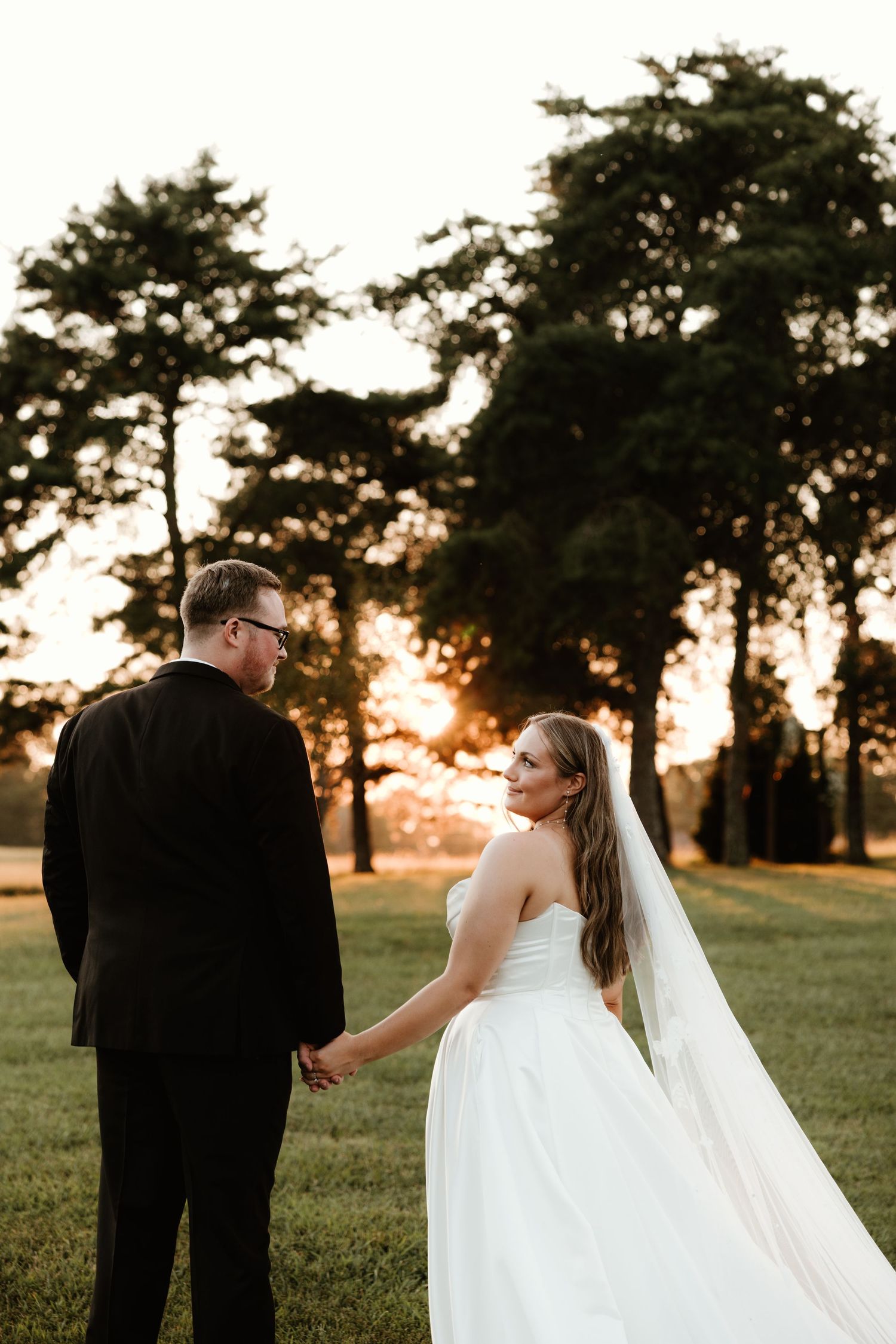 A couple shares an intimate sunset moment in a grassy field with pine trees silhouetted in the golden light.