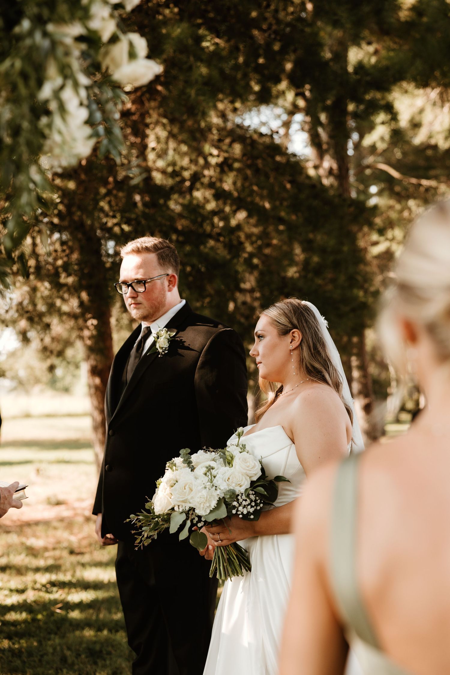 Outdoor wedding ceremony beneath trees with sunlight filtering through branches, featuring white bridal bouquet.