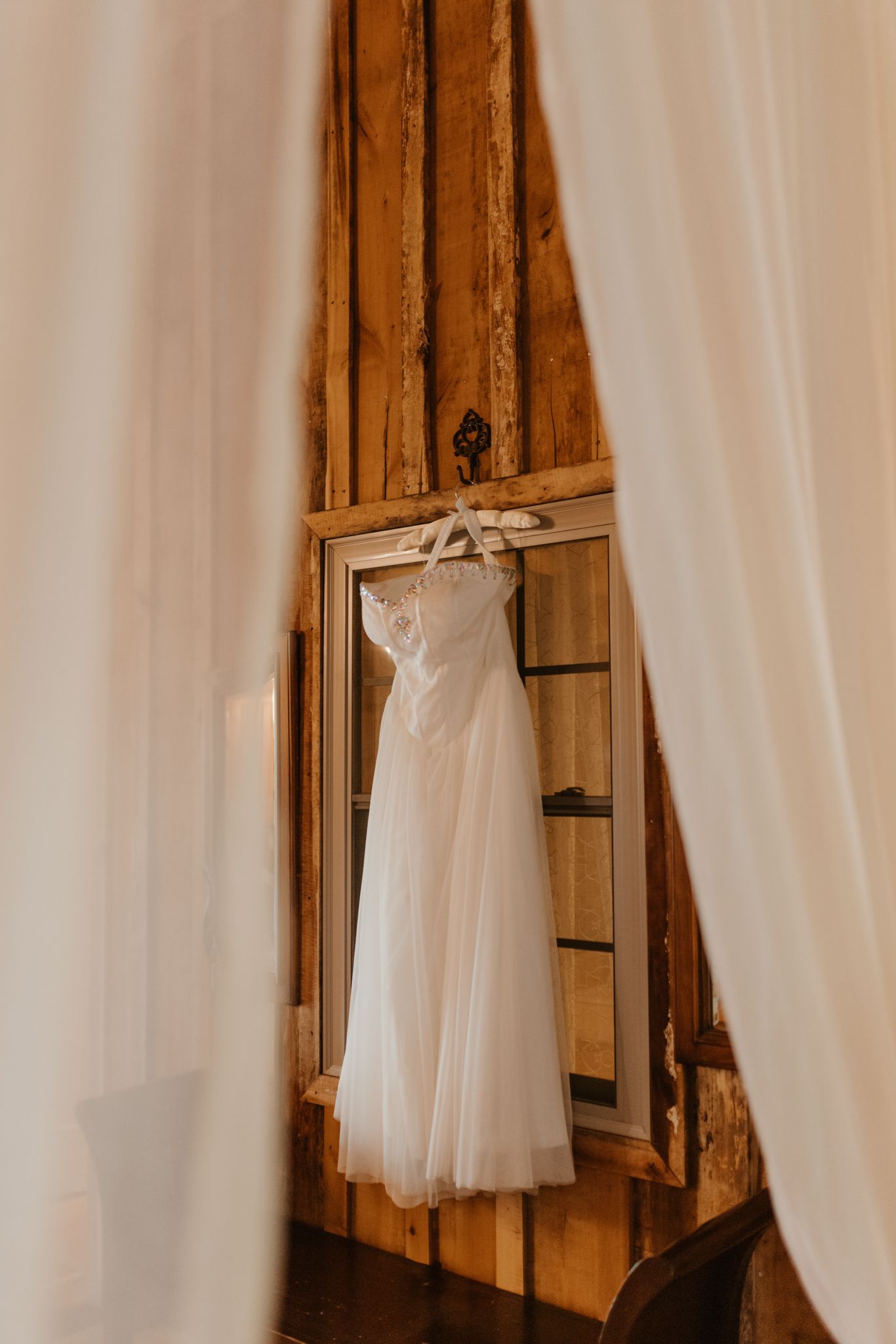 Elegant white wedding dress hanging on wooden wall between white curtains in rustic interior setting.
