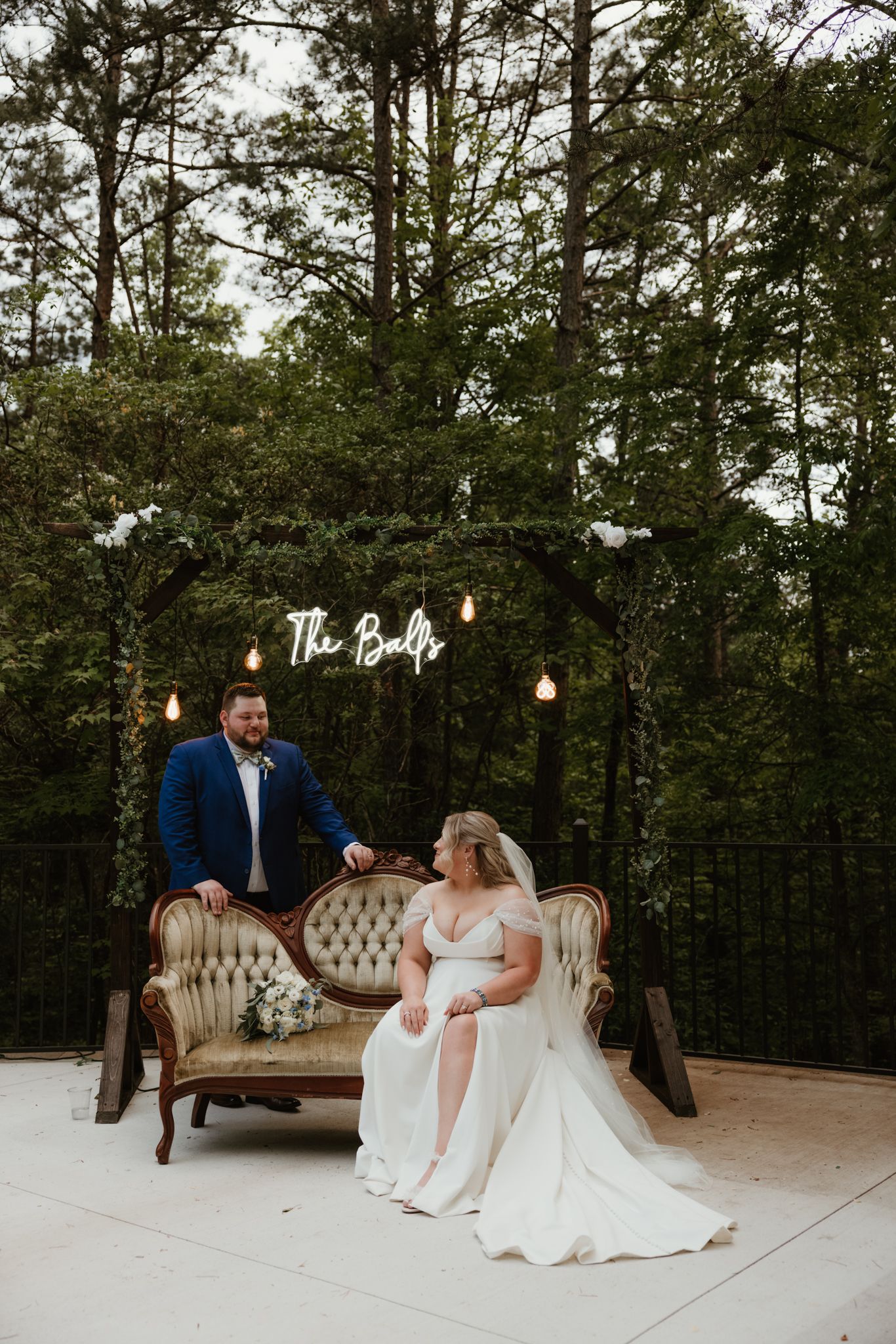 Wedding couple relaxes on vintage sofa under illuminated sign and forest backdrop.