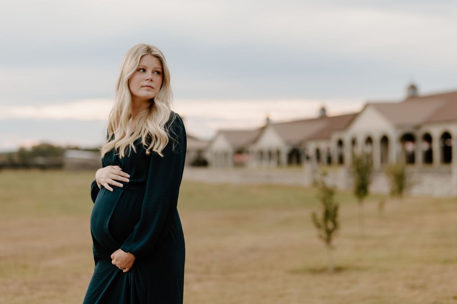 A person in a dark green dress stands in front of a rustic ranch building during golden hour.