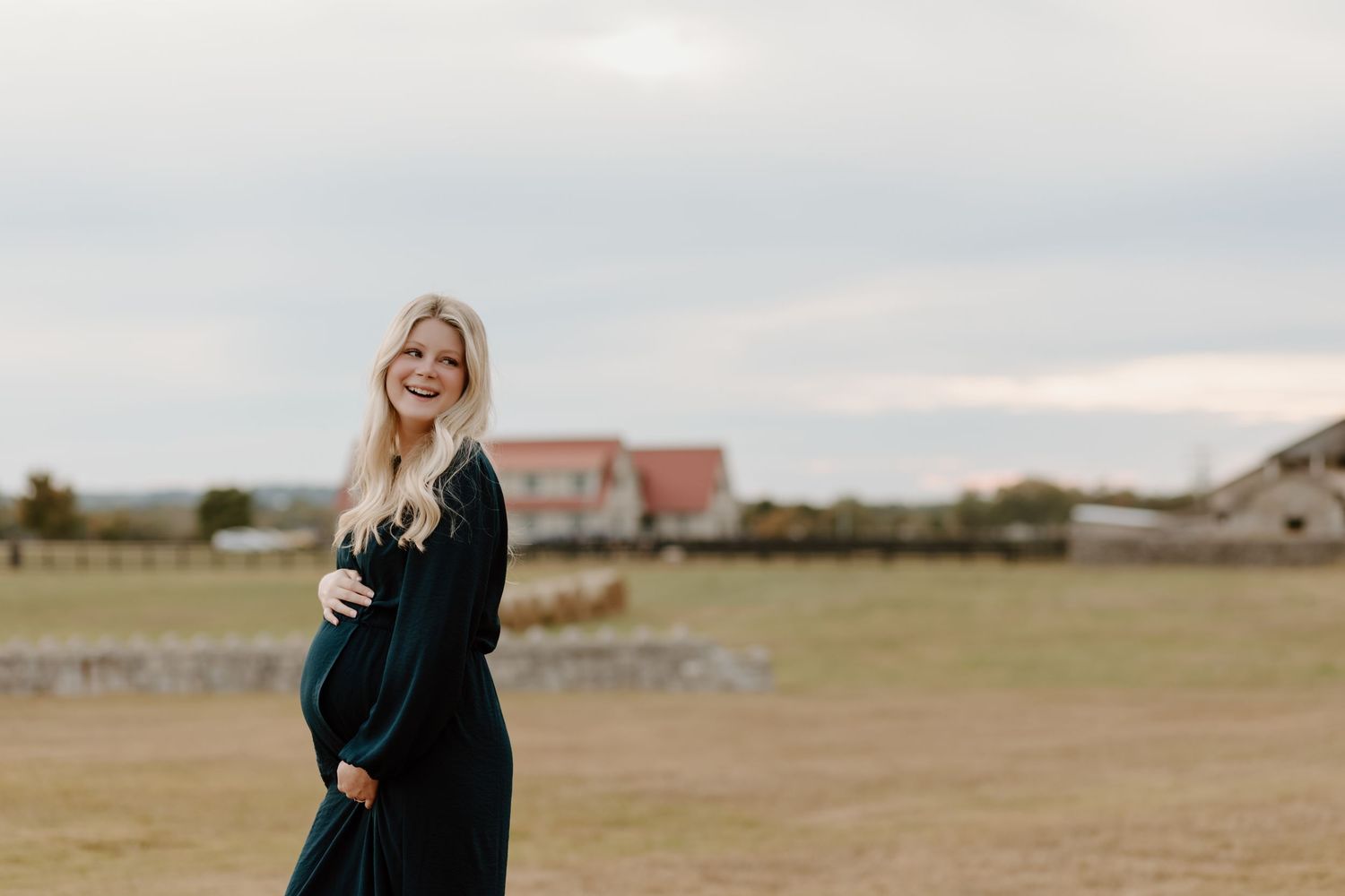 A pregnant woman in a navy dress smiling while standing in an open field.