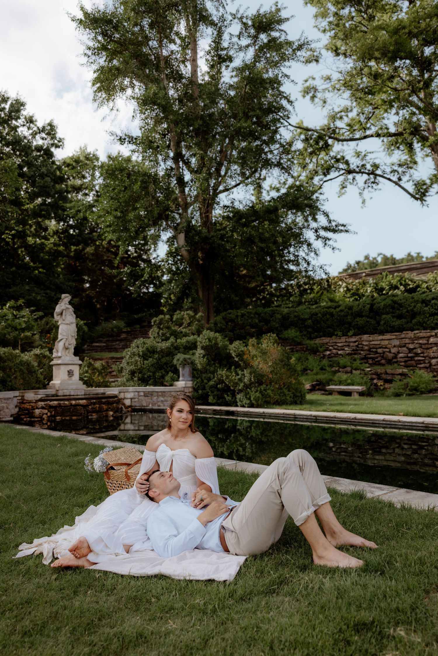 Couple relaxing on white blanket during romantic garden picnic near stone fountain and classical statue.