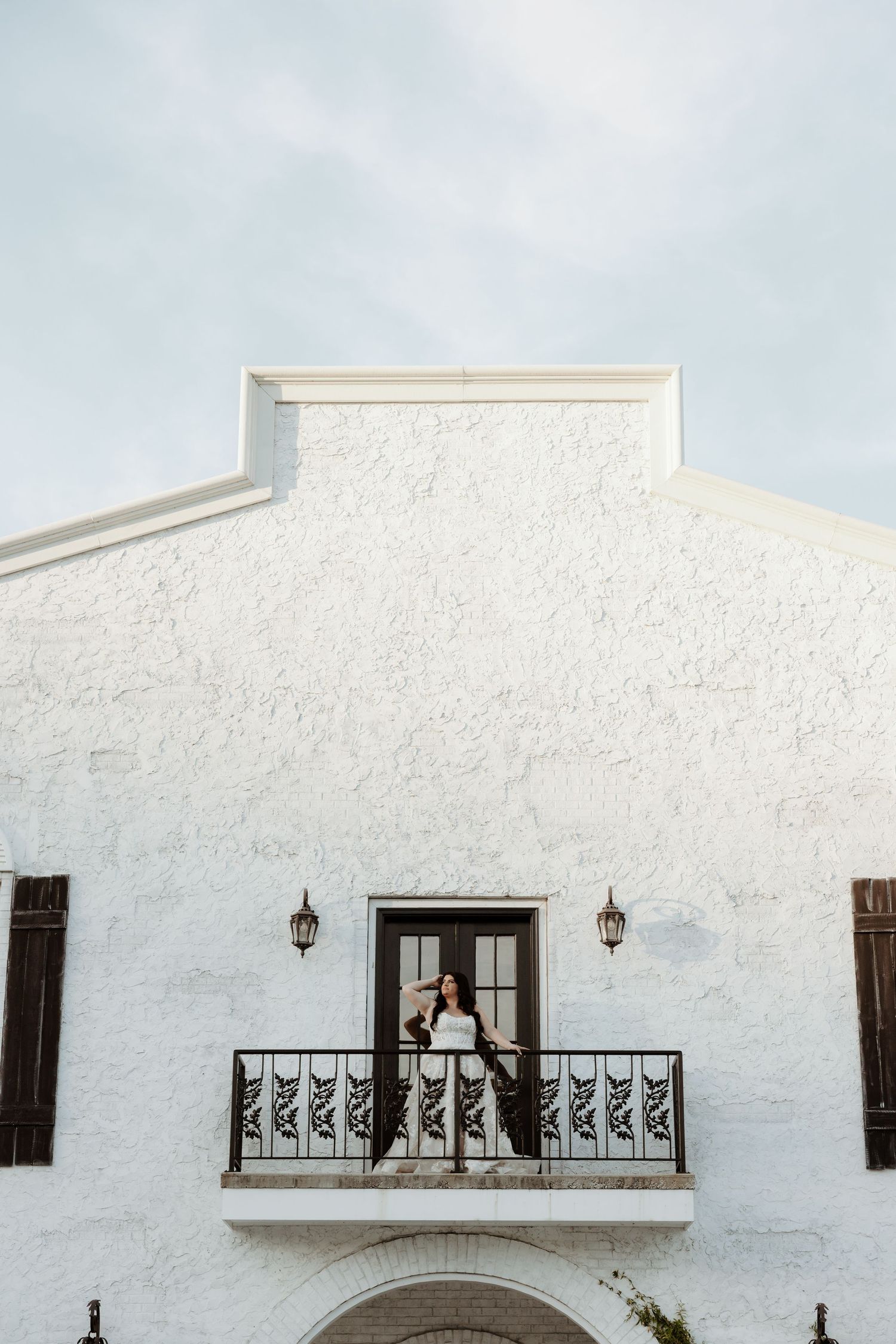 A bride in a black and white gown stands on an ornate balcony of a white stucco building with dark shutters.
