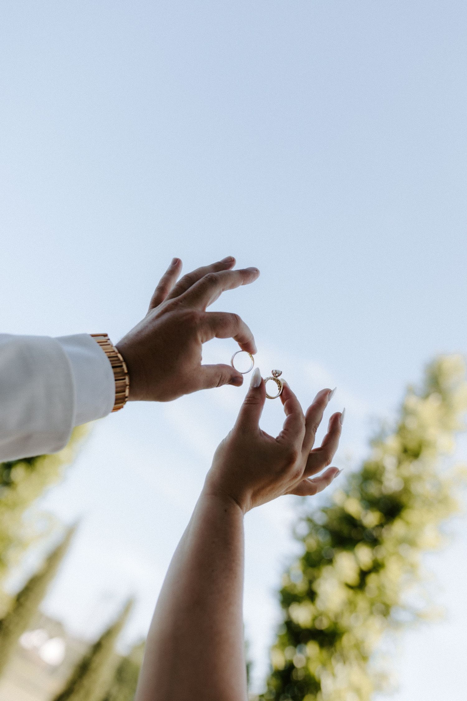 Two hands exchange wedding rings against a clear blue sky, one wearing a gold bracelet.