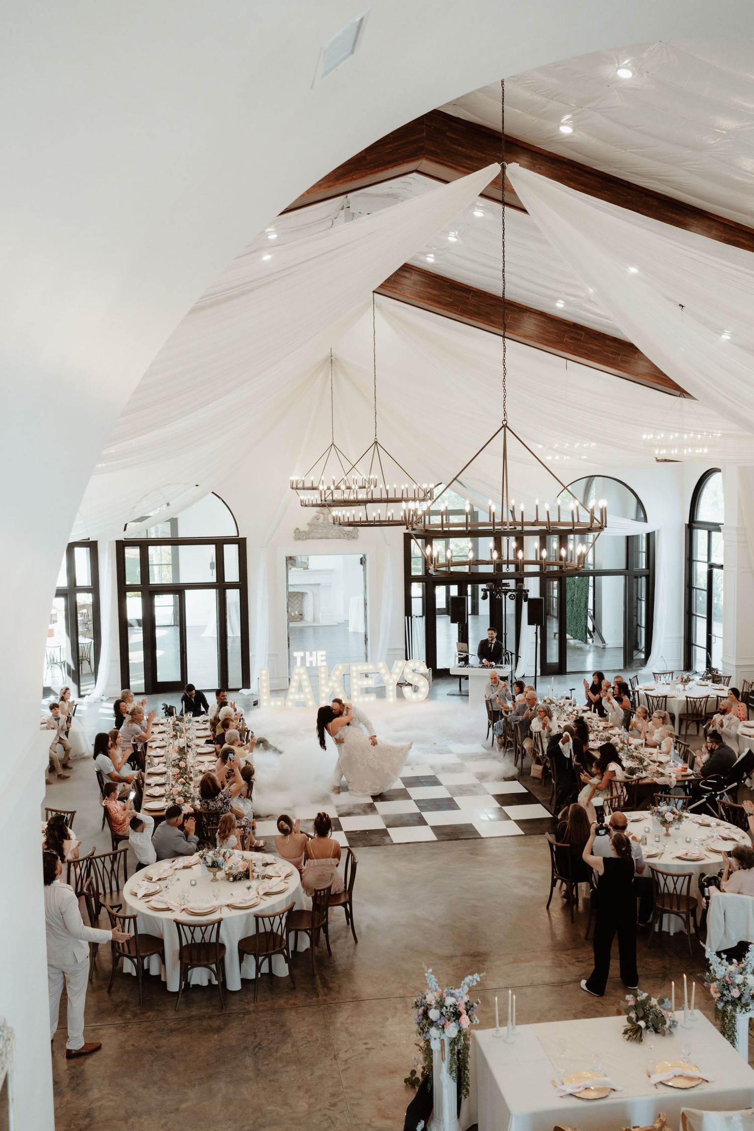 Stunning wedding venue with vaulted ceilings, white draping, chandeliers, and checkered dance floor with guests dining.