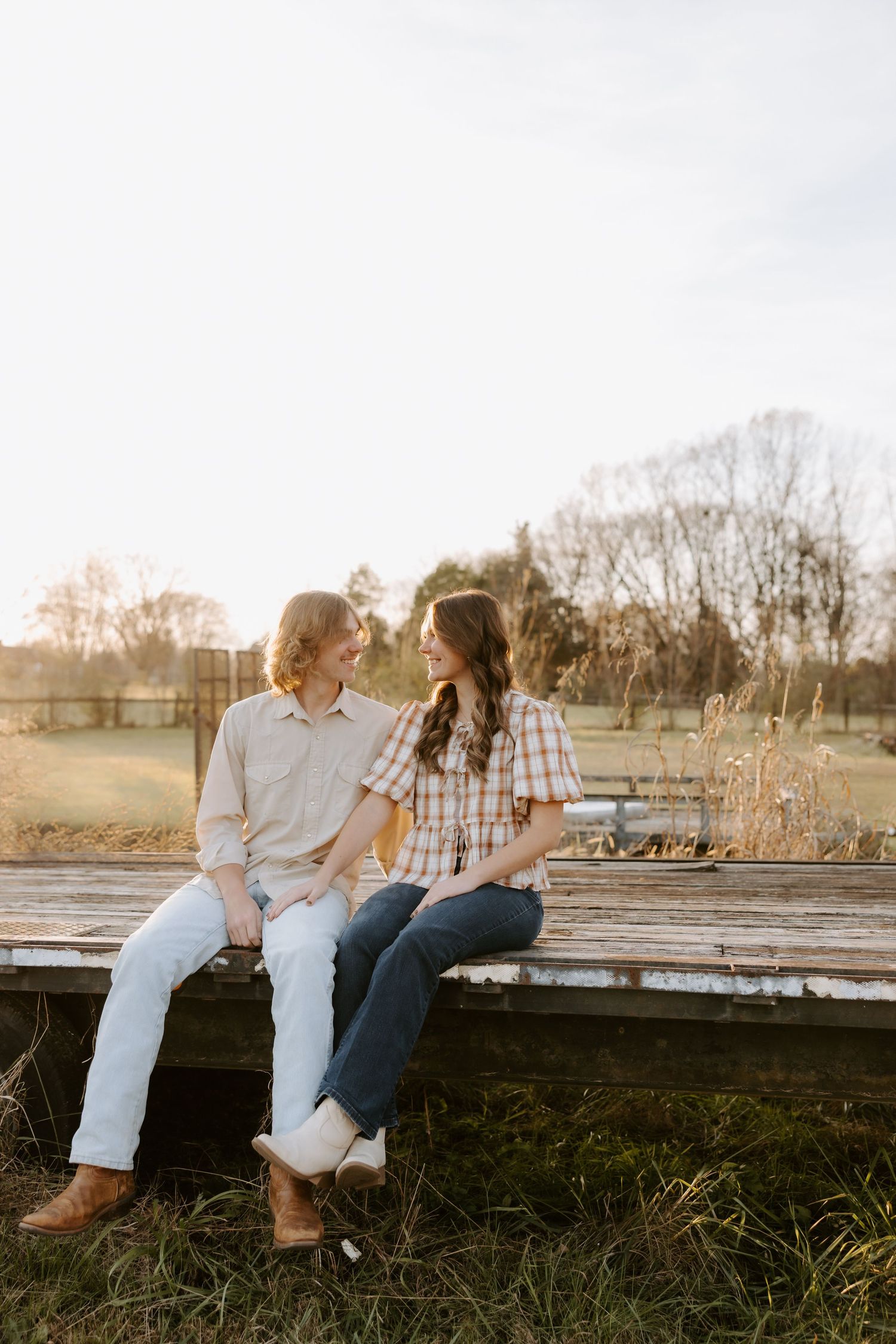 A couple sits on a rustic wooden bench in a golden hour farm setting, gazing at each other in matching plaid shirts and jeans.