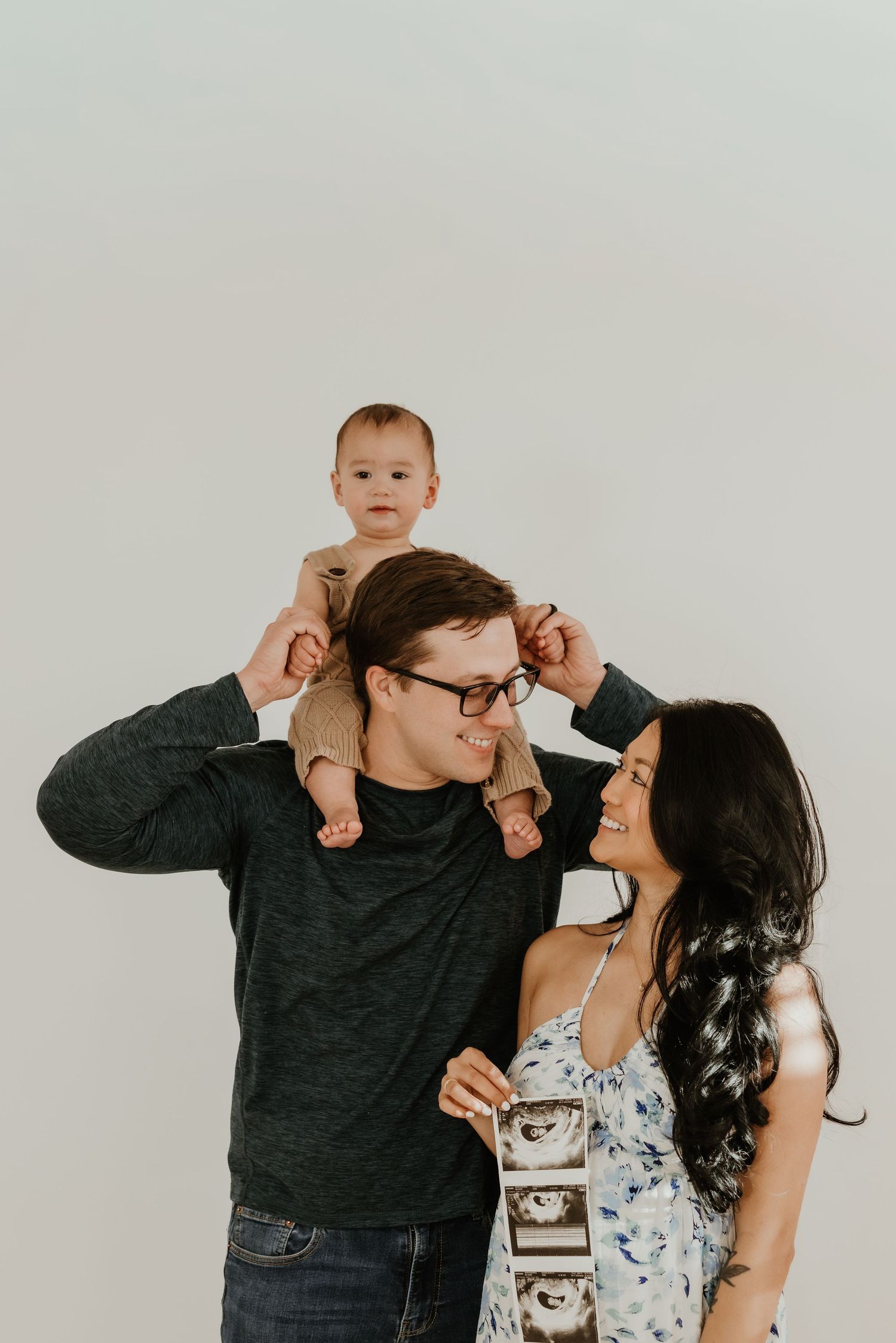 Happy family portrait with dad holding baby on shoulders, mom showing ultrasound photo, announcing second pregnancy.