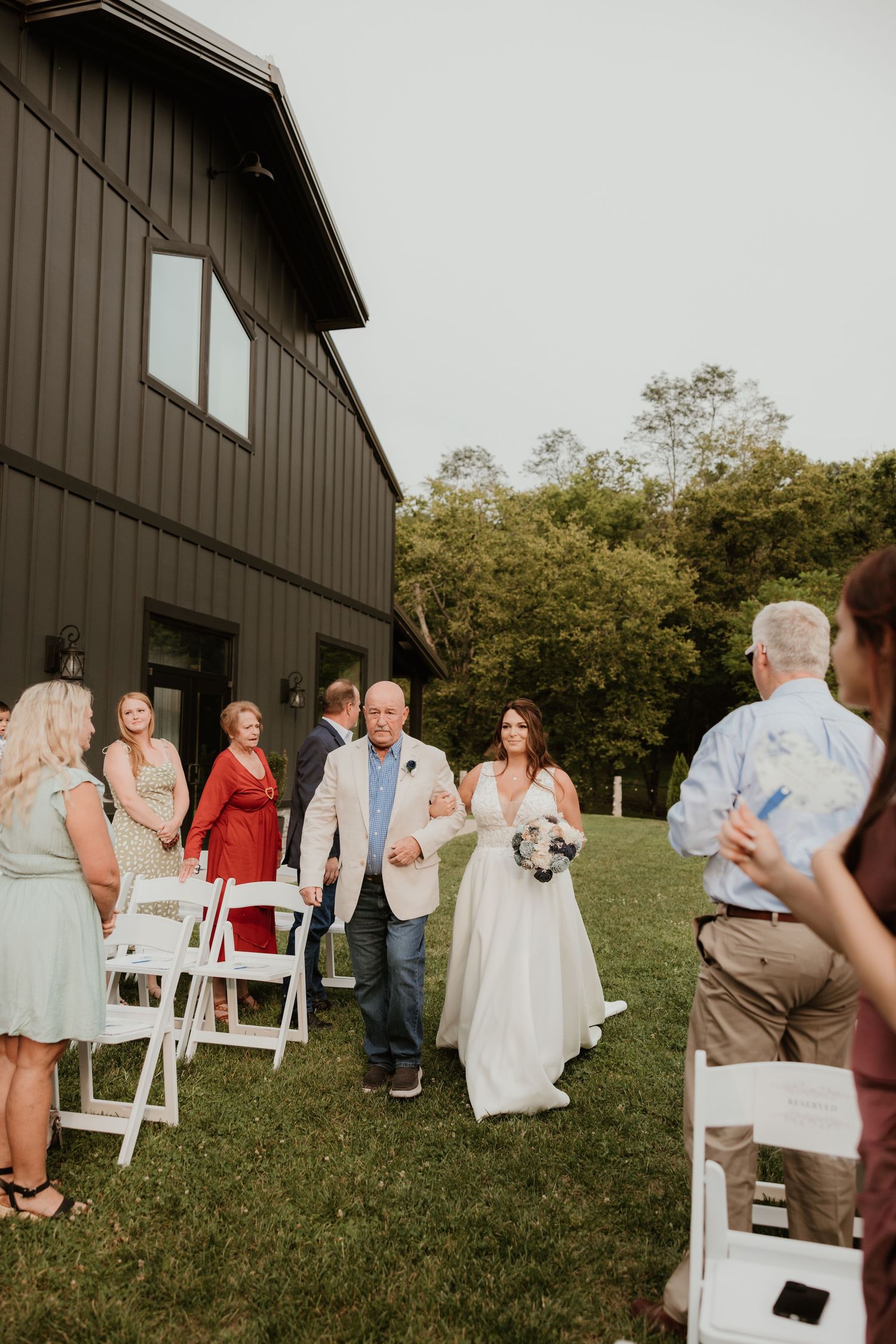 Wedding guests gather on lawn chairs outside a rustic barn venue during an outdoor ceremony.