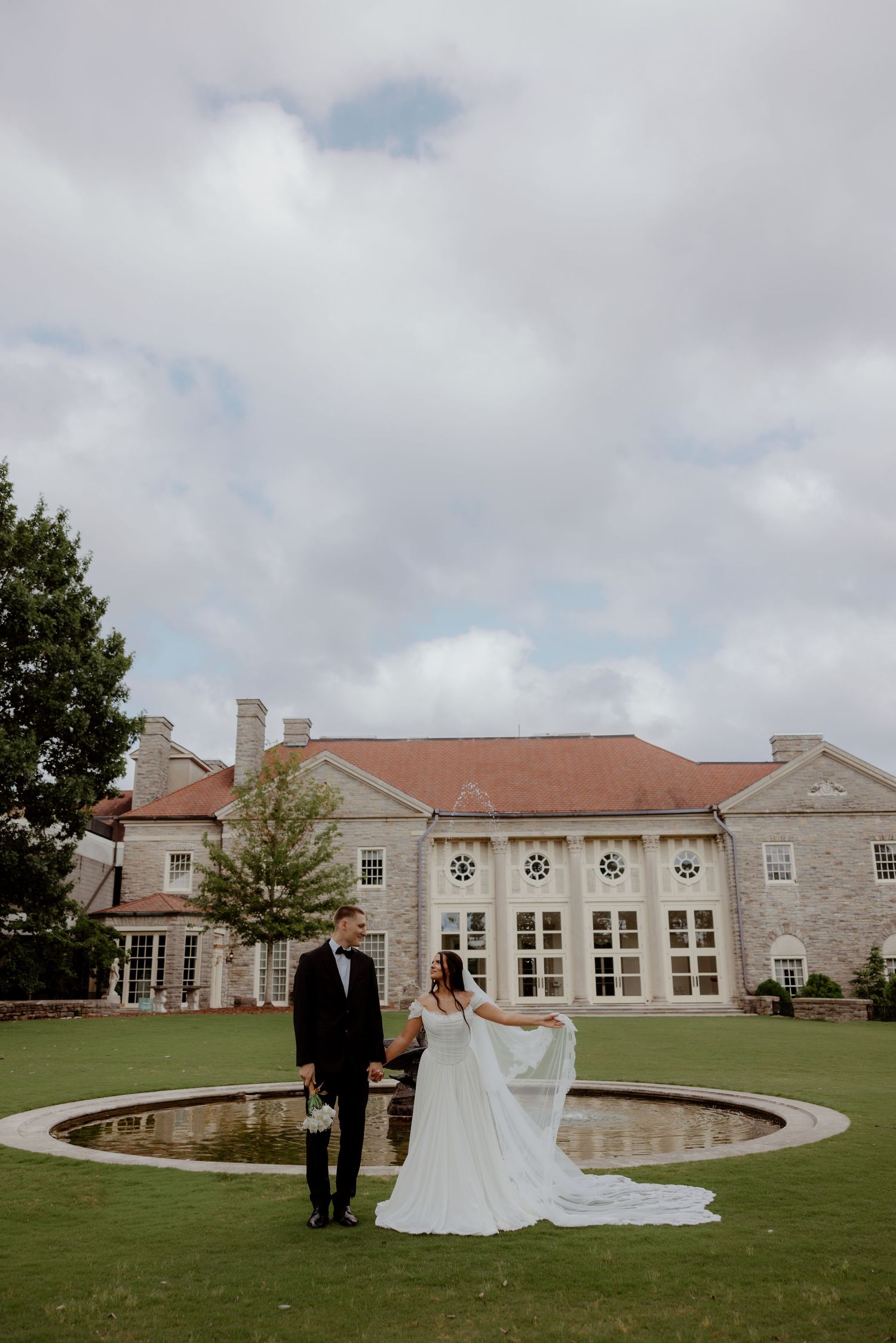 Bride and groom hold hands on manicured lawn before elegant stone mansion wedding venue under cloudy sky.