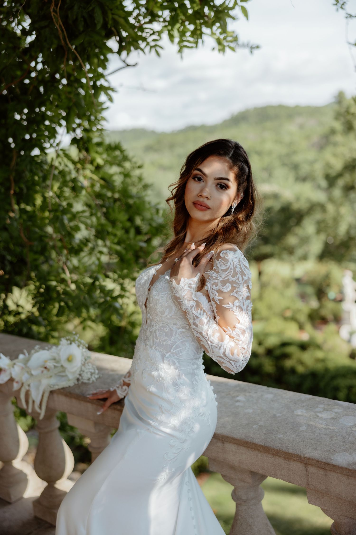 Bride in lace long-sleeve mermaid wedding dress poses on stone balcony with lush green hills in background.