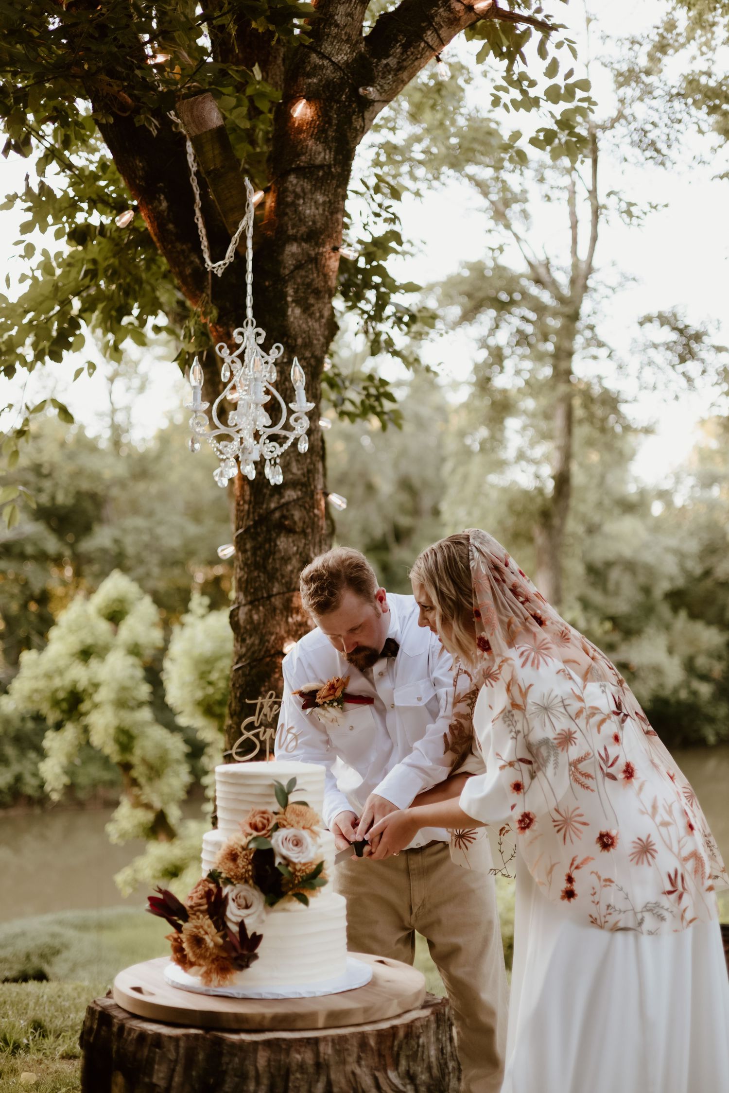 A couple and their small dog share a sweet moment while cutting a cake at their outdoor celebration in the woods.