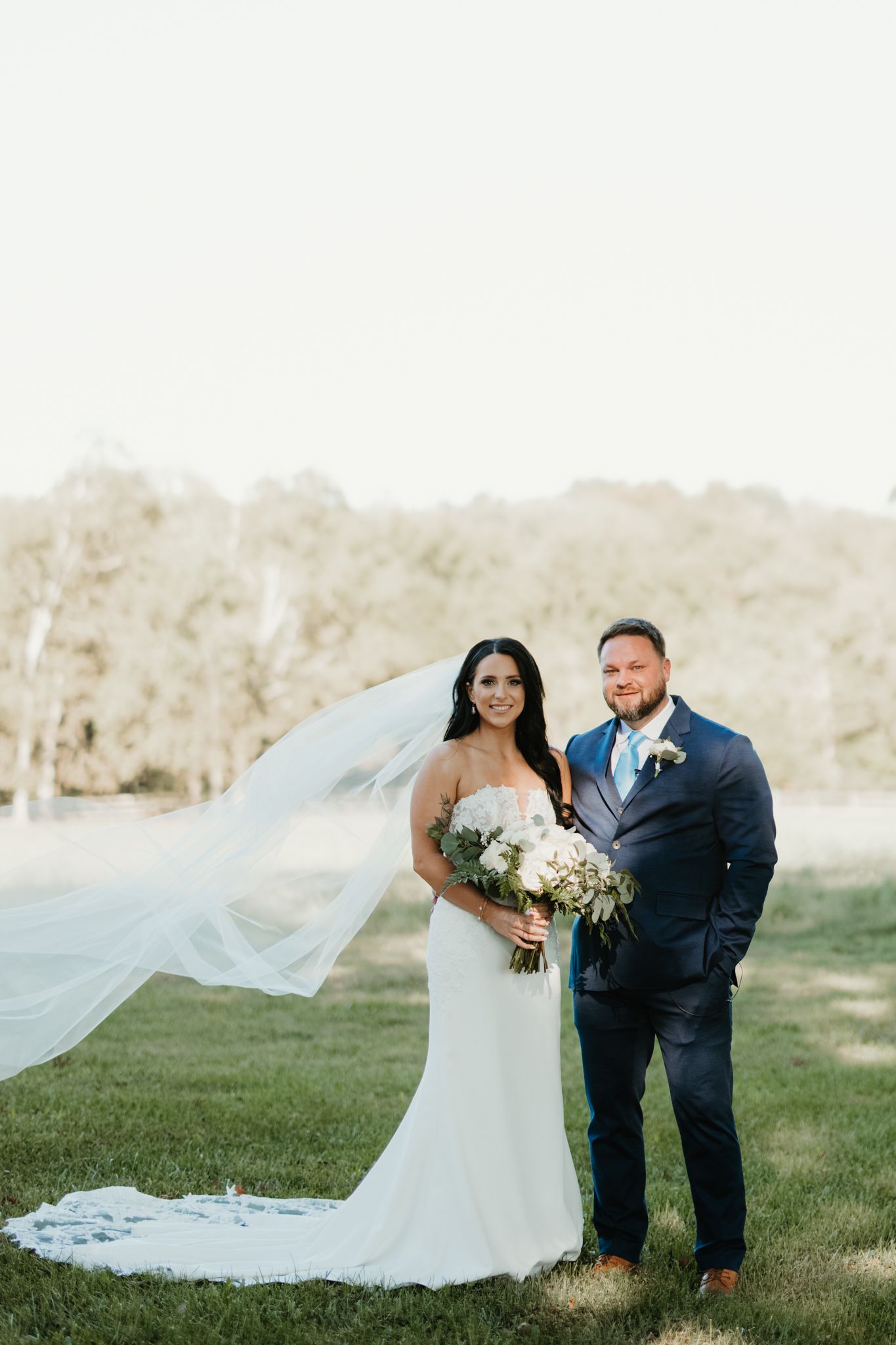 Wedding couple posing outdoors with long flowing veil catching wind in natural setting.