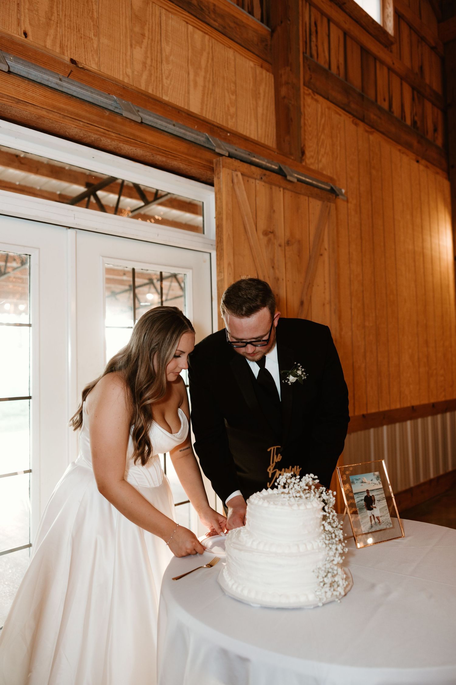 A wedding couple stands at a white table cutting their wedding cake together in a rustic wooden barn venue.