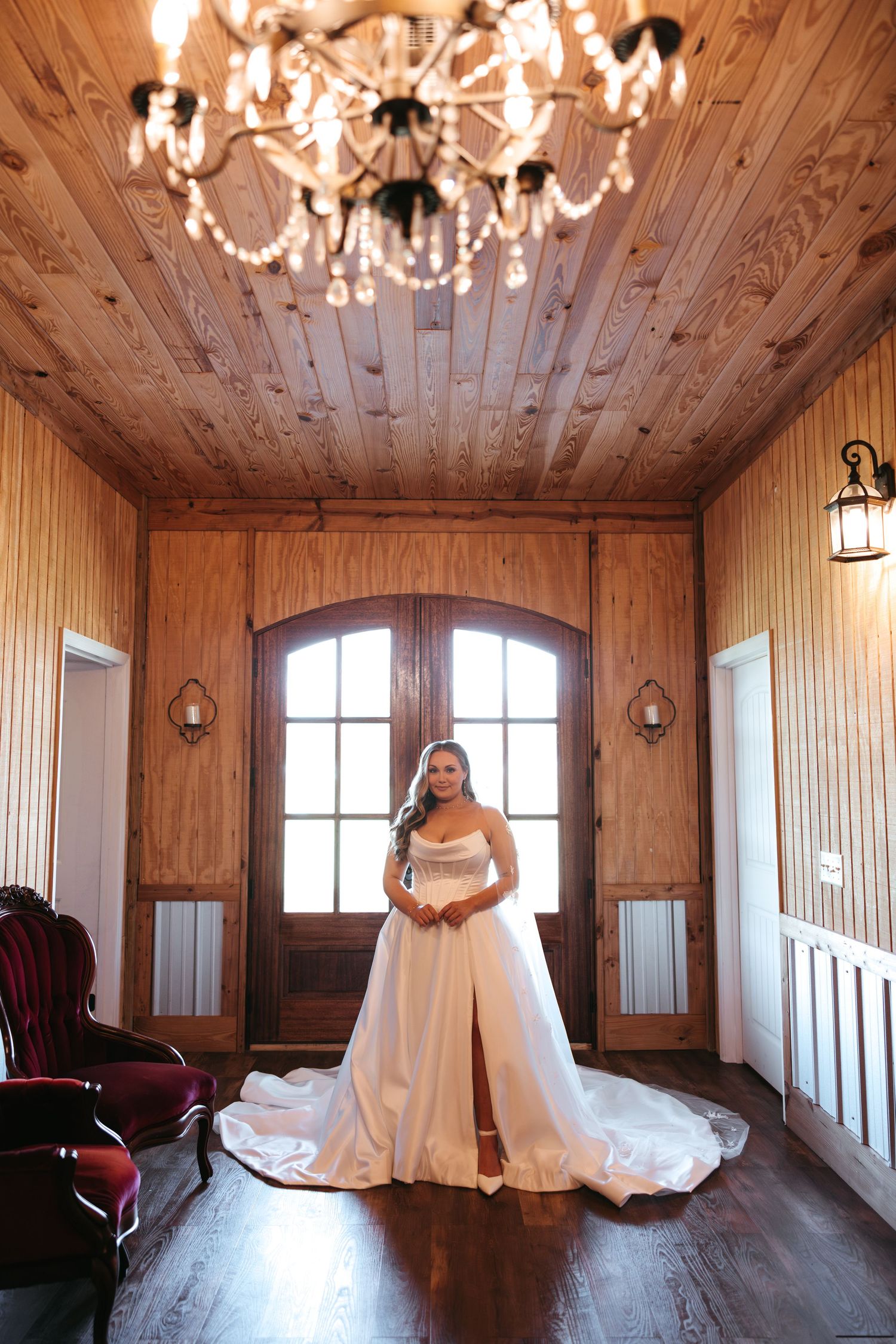 A stunning elegant bridal photograph with a flowing white gown in a rustic wooden room with a sparkling chandelier.