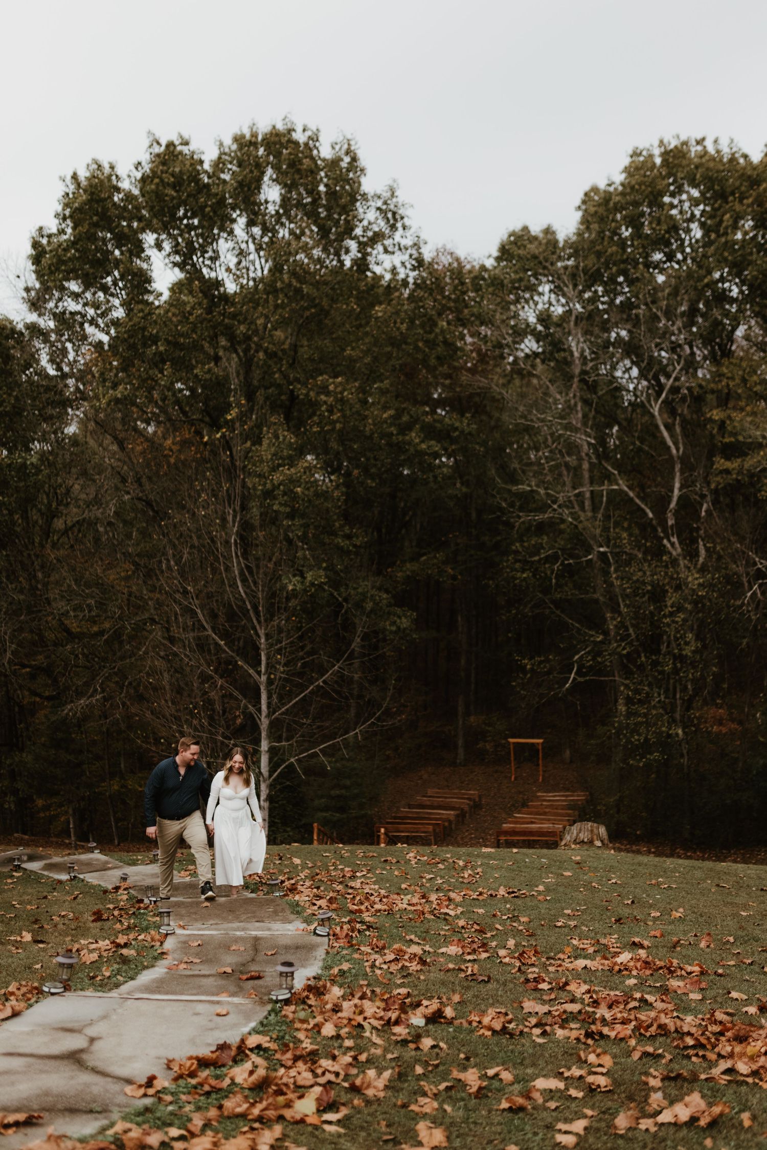 A couple walks along a stone path surrounded by autumn leaves and tall trees.