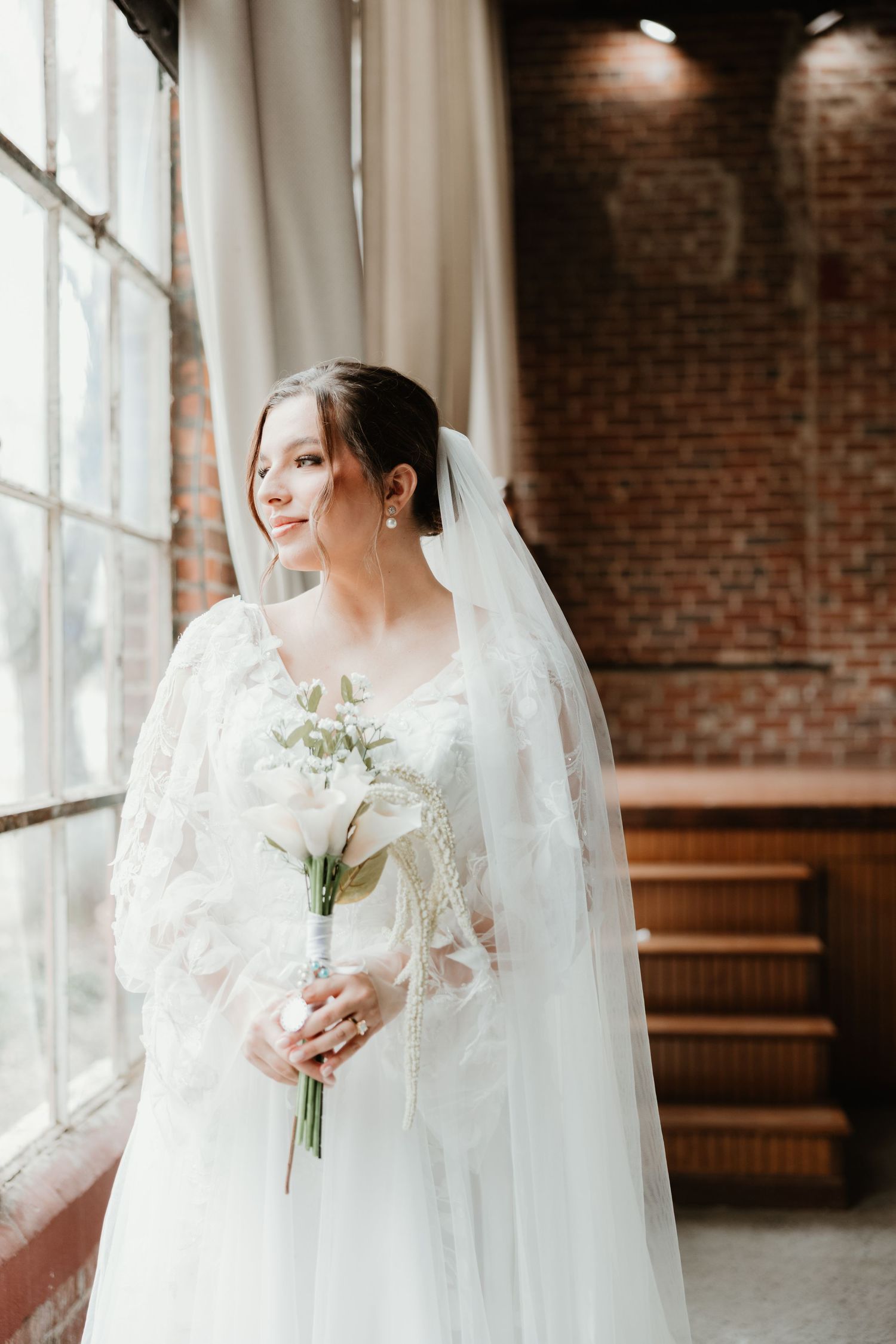 Bride in white lace gown holds bouquet while standing by sunny window.