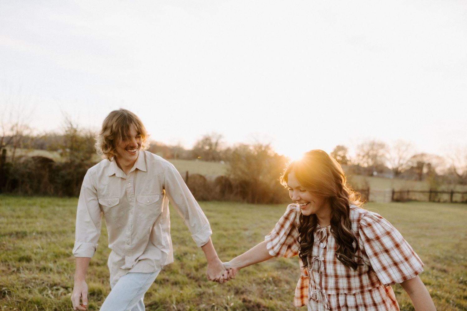 A couple holds hands and laughs together in a sunlit field during golden hour.
