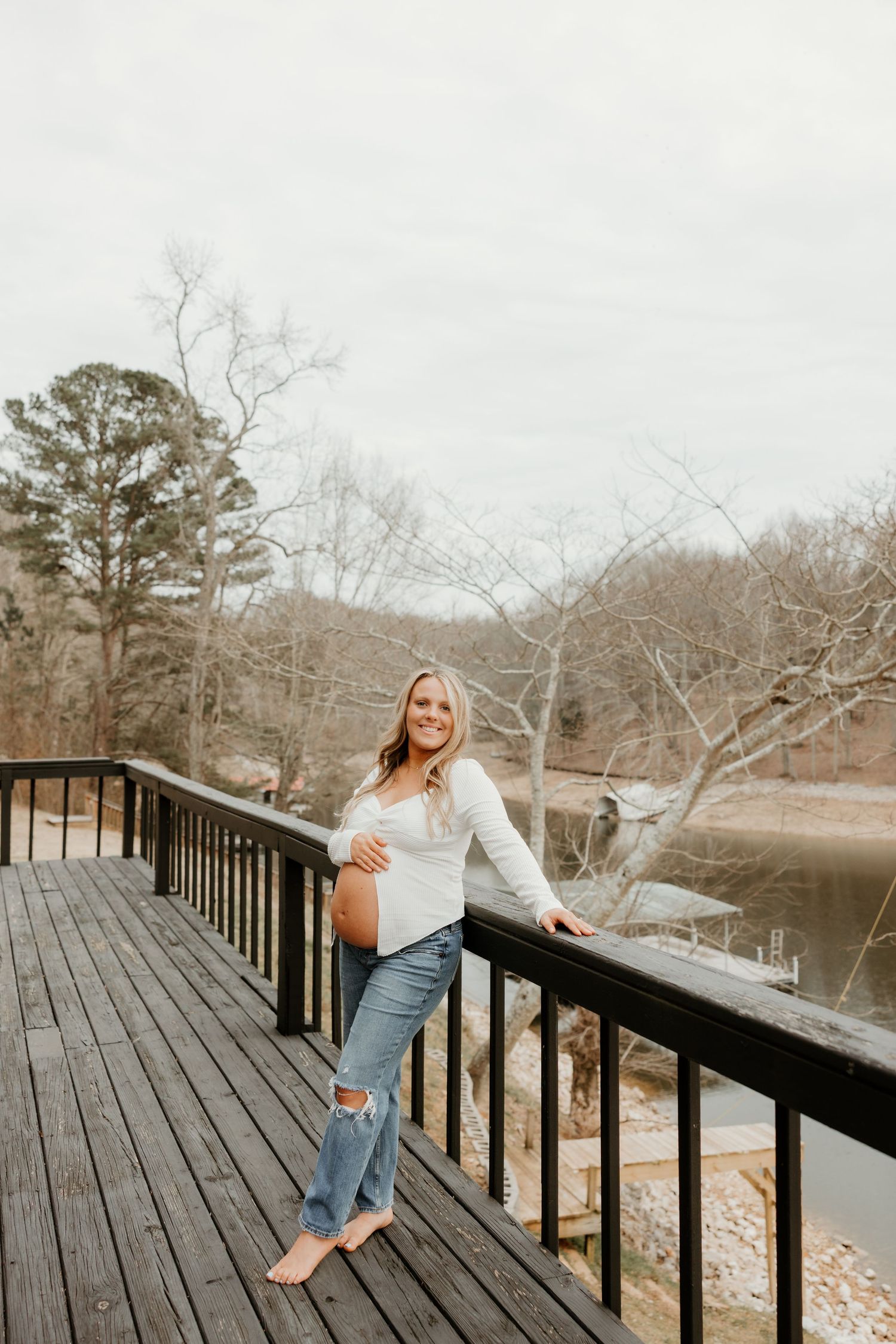 Smiling pregnant woman in white top and ripped jeans poses on wooden deck overlooking a serene lake surrounded by trees.