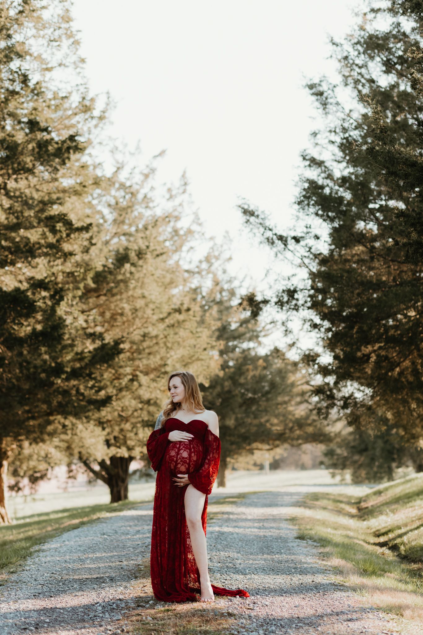 Figure in a burgundy dress poses elegantly on a tree-lined gravel path at golden hour.