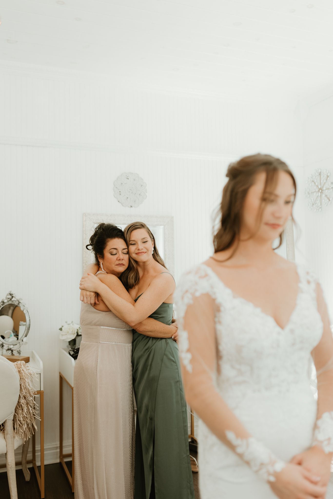 Emotional candid moment at a bridal fitting shows intimate embrace in background while bride stands in lace gown.
