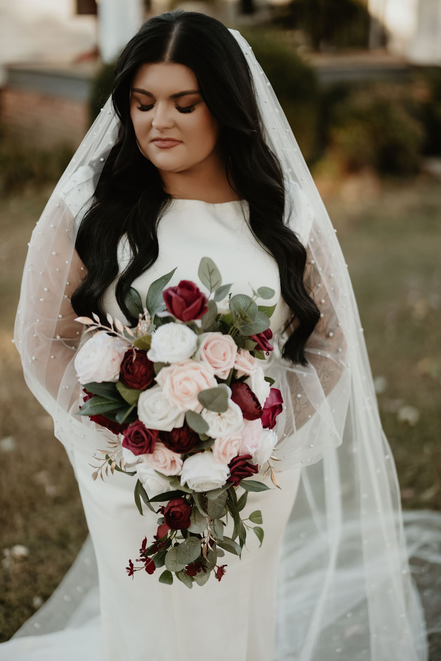 A wedding bouquet of burgundy and blush roses with eucalyptus held against a white gown in natural light.