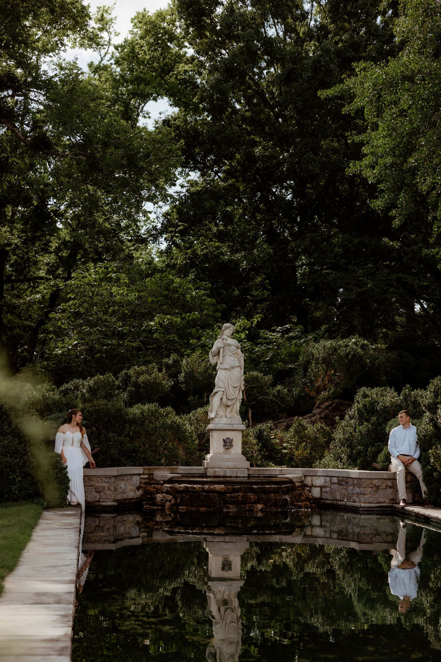 Couple poses beside a stone statue fountain reflecting in a pool, surrounded by lush green trees in an elegant garden.