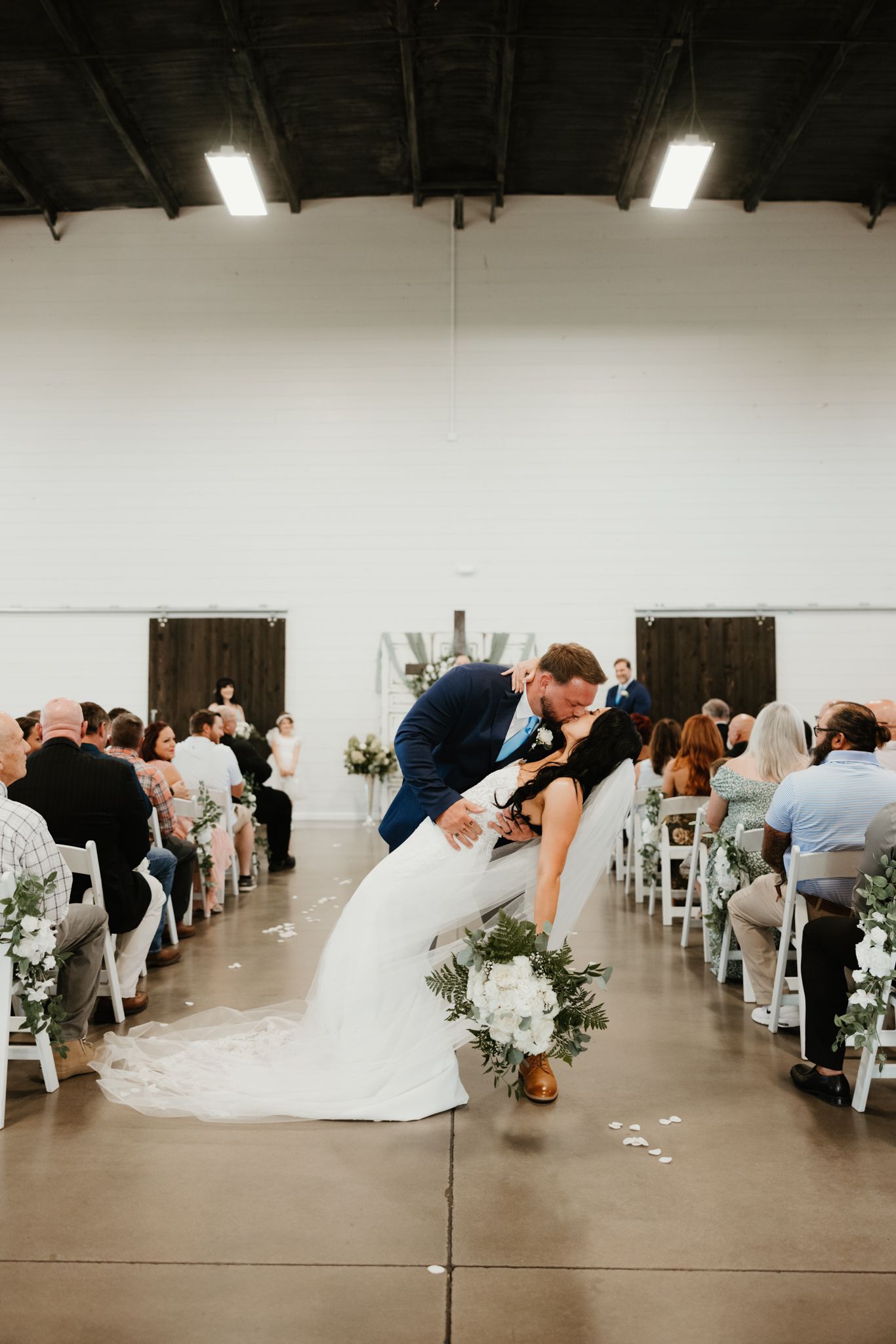 Wedding ceremony in a white industrial venue with guests seated in rows and a romantic dip at the aisle.